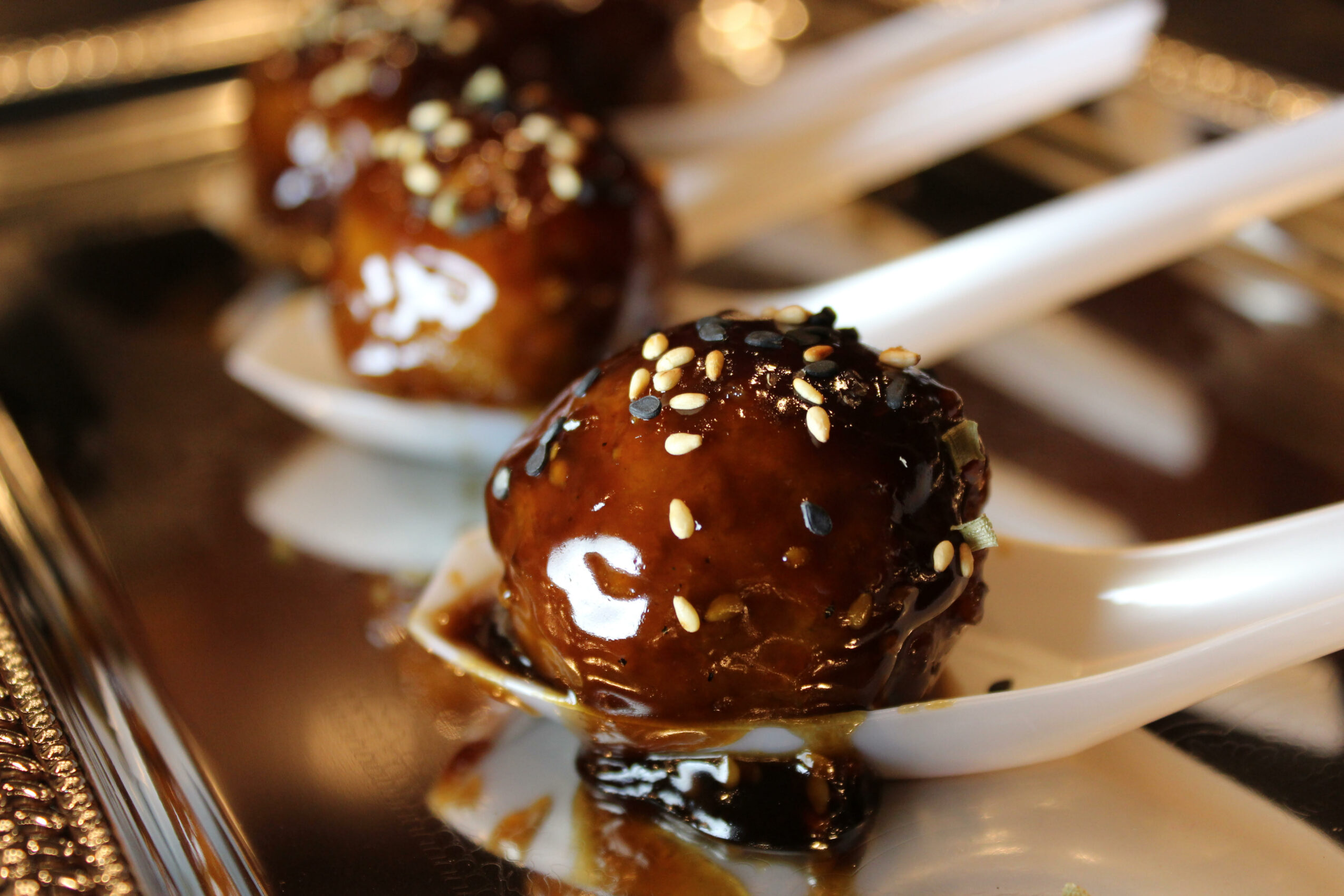 Close-up of a glazed meatball with sesame seeds on a white spoon, with another meatball in the background.