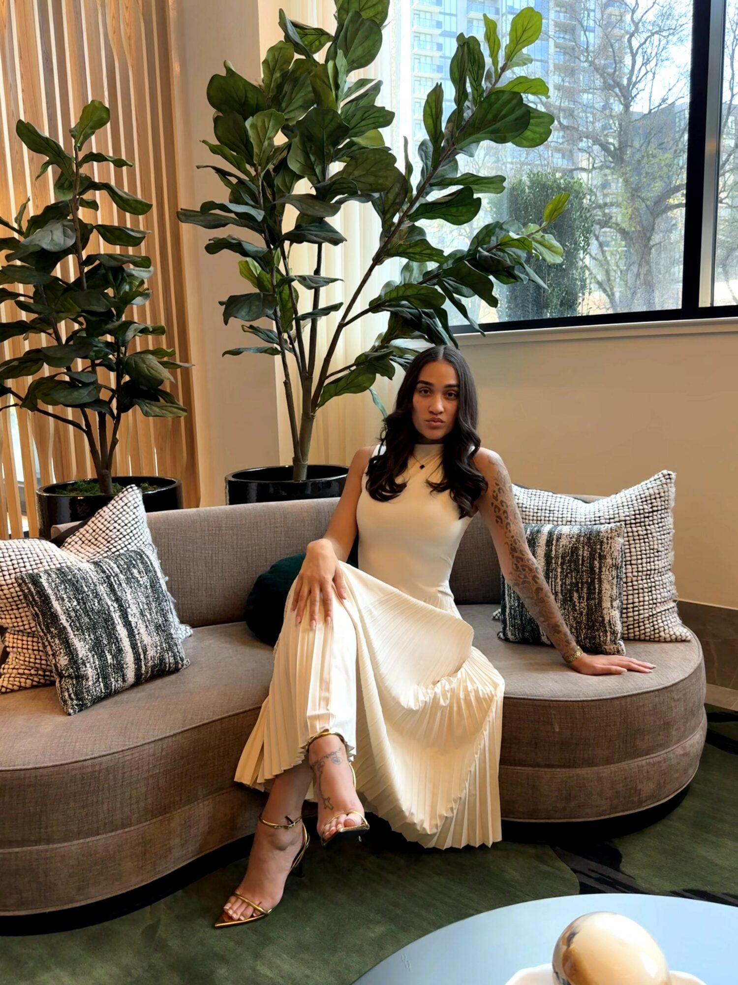 Woman in a white dress sitting on a curved sofa with patterned pillows, large green plant behind her, window in background.