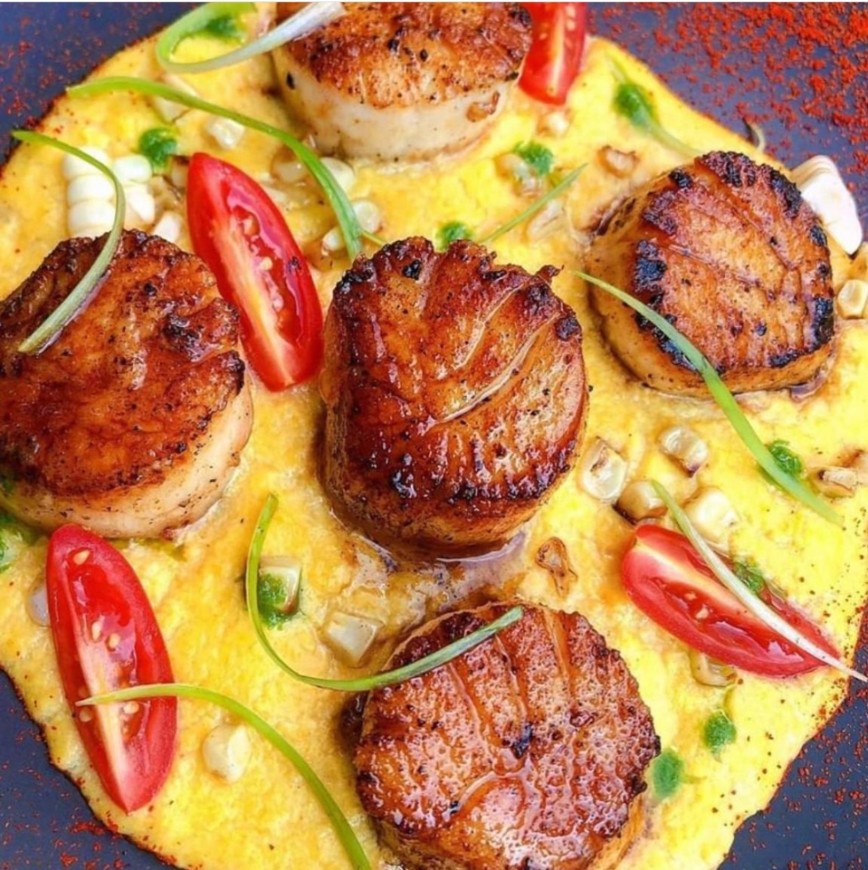 Seared scallops on a yellow sauce with cherry tomatoes and green garnish, garnished with chopped nuts or garlic, on a black plate.