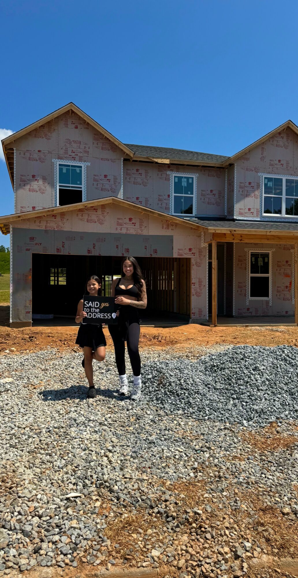 Two children stand in front of a house under construction, with a clear blue sky above.