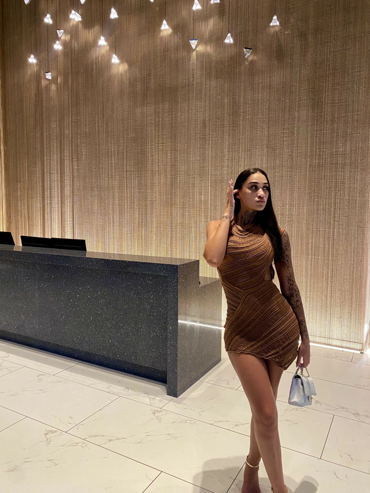 A woman with long dark hair in a brown dress stands in a modern interior, holding a small bag, with a textured wall and ceiling lights behind her.