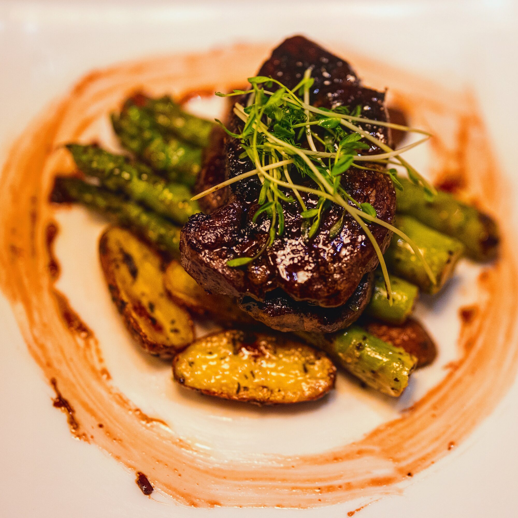 Steak with grilled vegetables and microgreens on a plate with sauce swirl, close-up view.