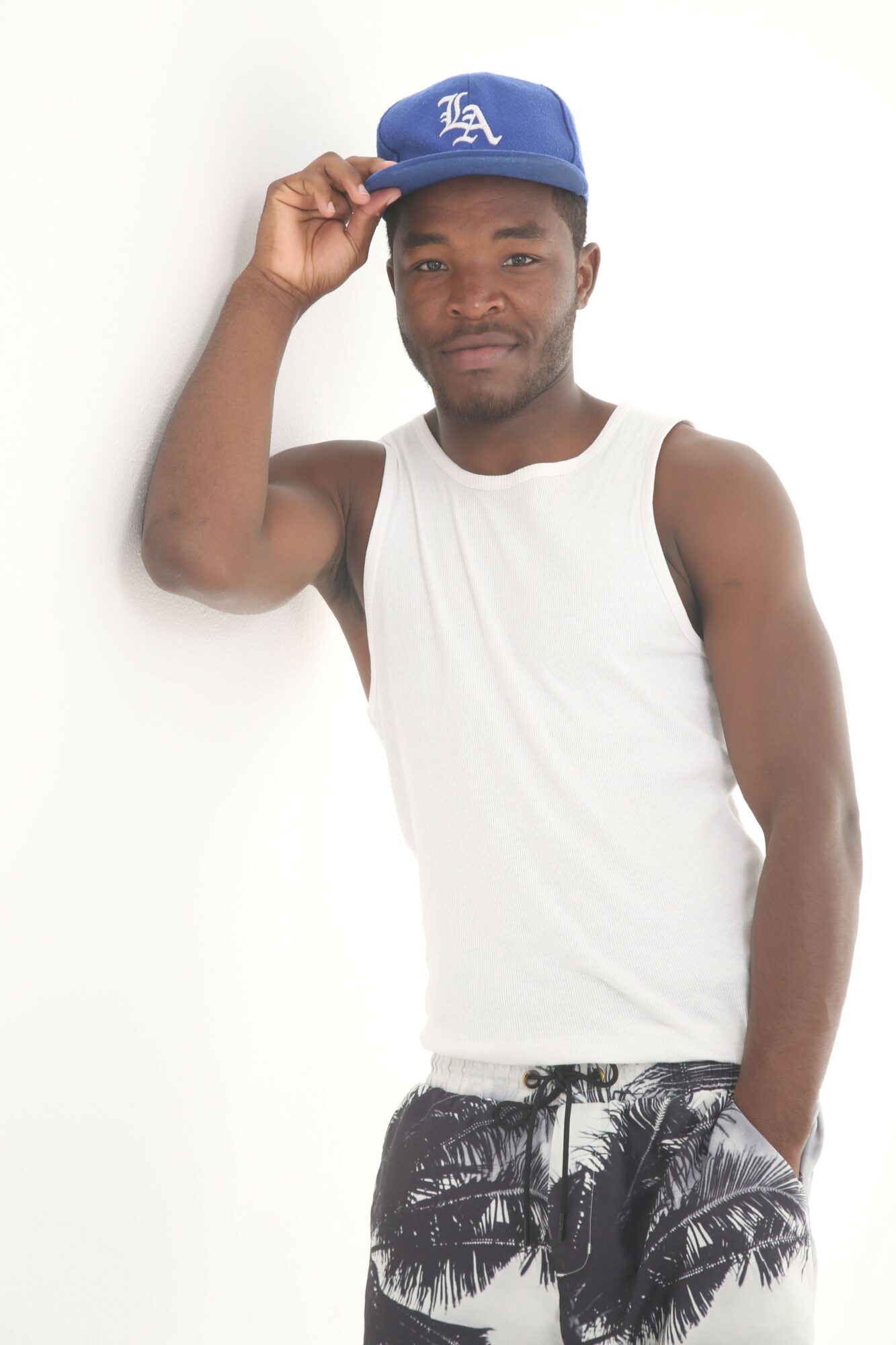 Man wearing a blue cap and white tank top, holding the cap's brim, standing against a plain white background.