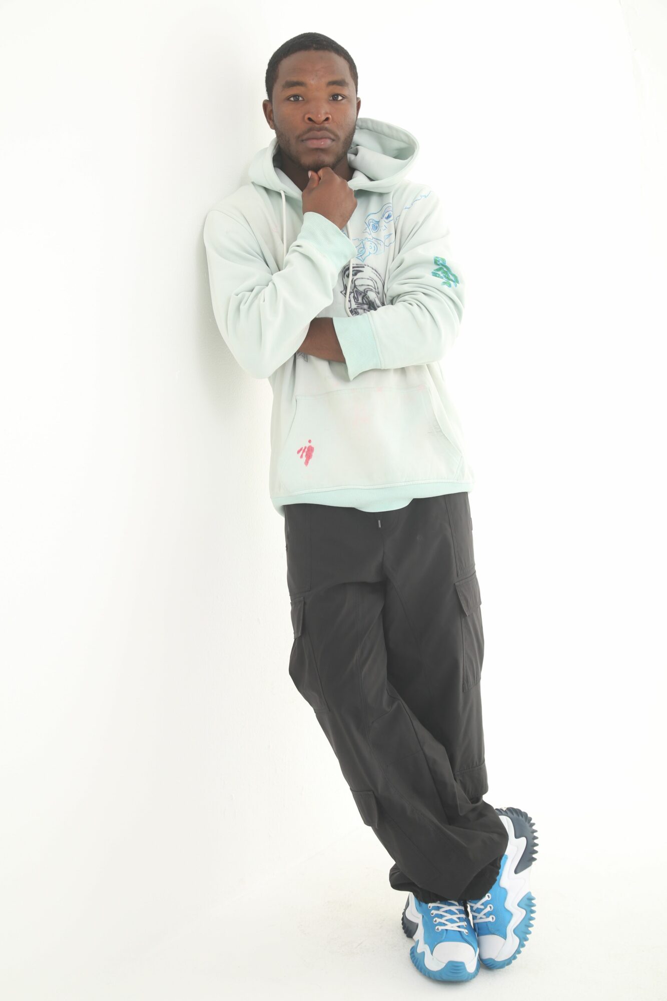 Person standing with arms crossed, wearing a white hoodie, black pants, and blue sneakers, against a white background.