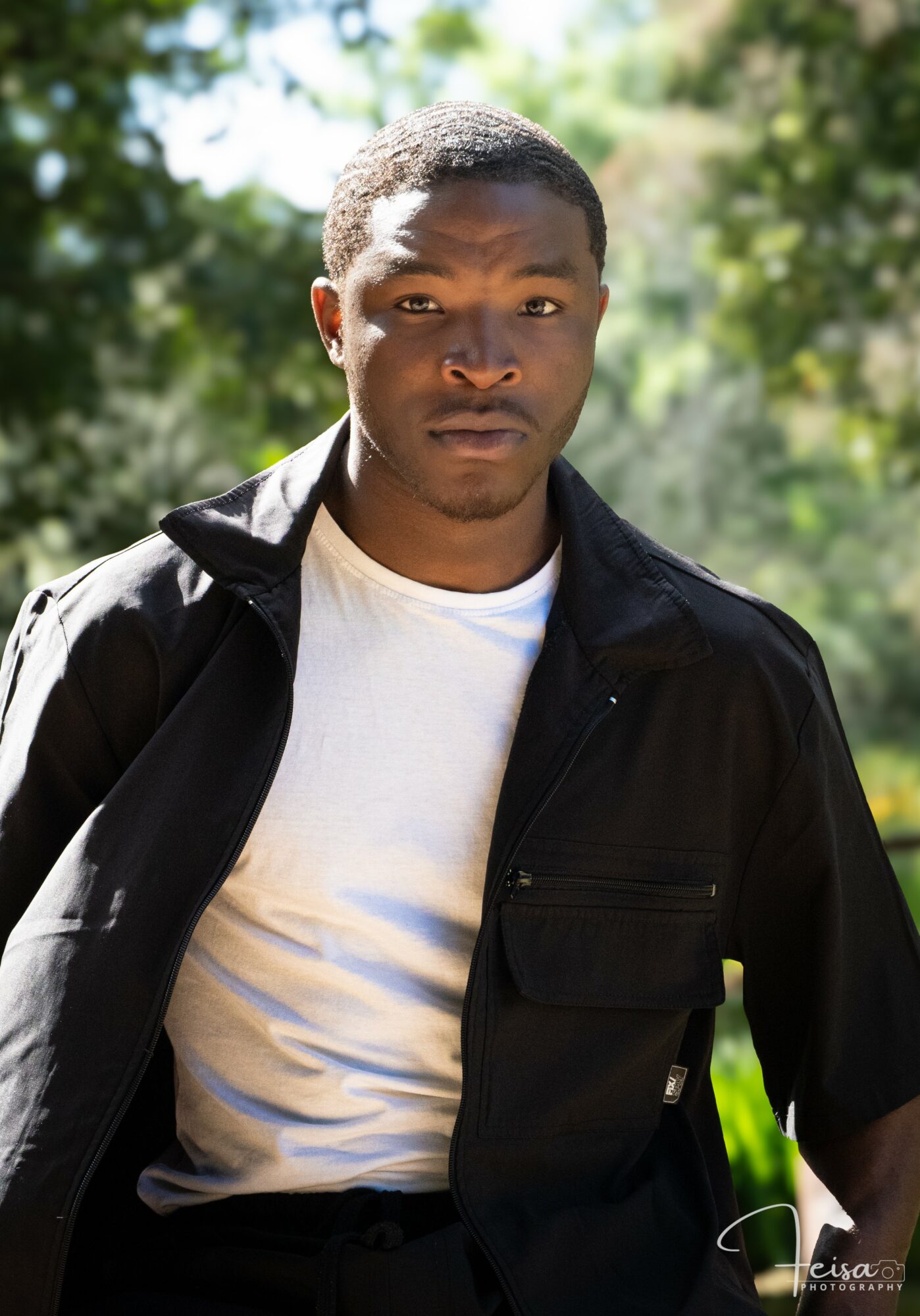 Young man outdoors wearing a white shirt and black jacket, with trees and sunlight in the background.