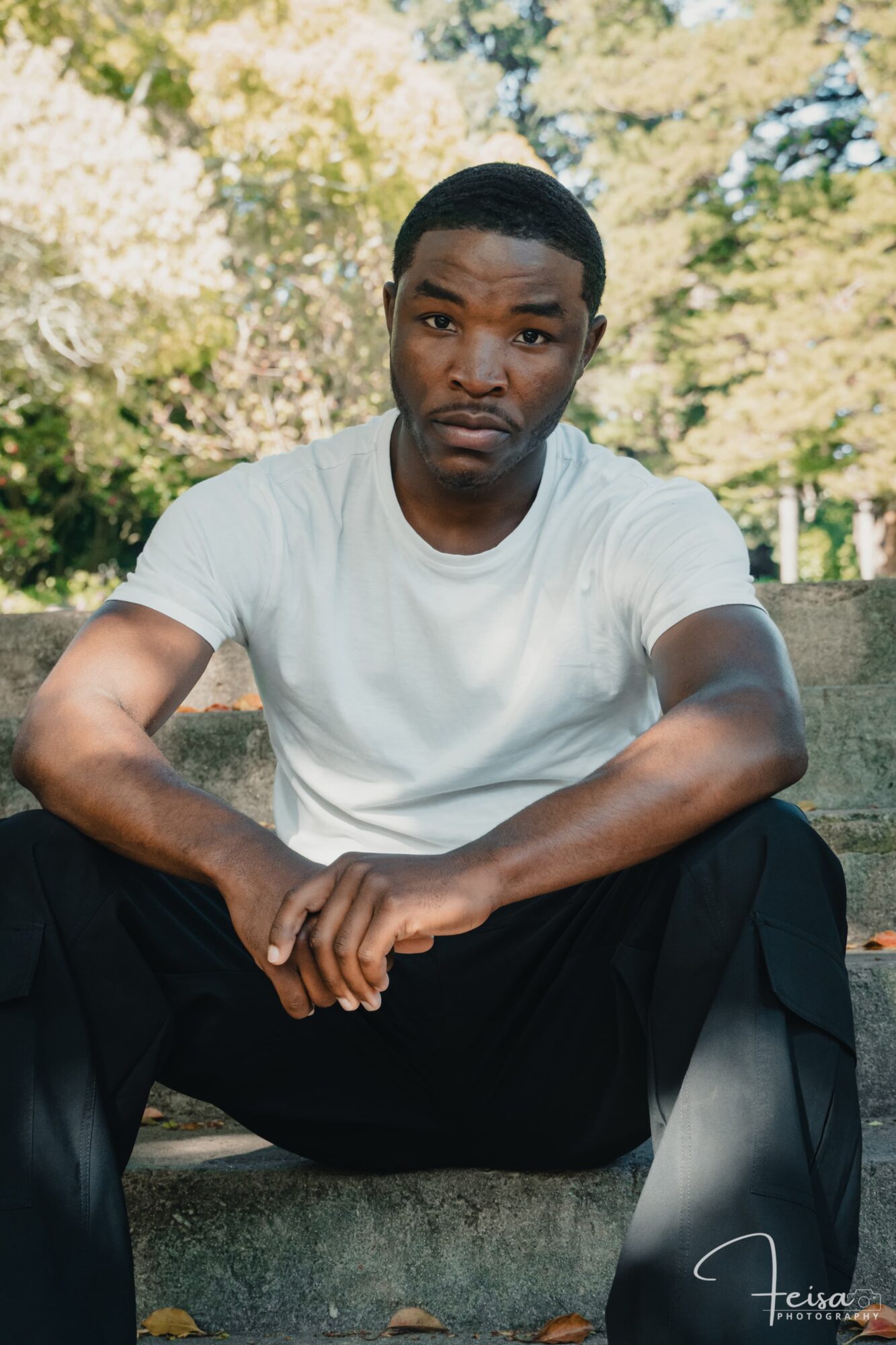 Man sitting outdoors on a concrete surface, wearing a white t-shirt, with trees and sunlight in the background.