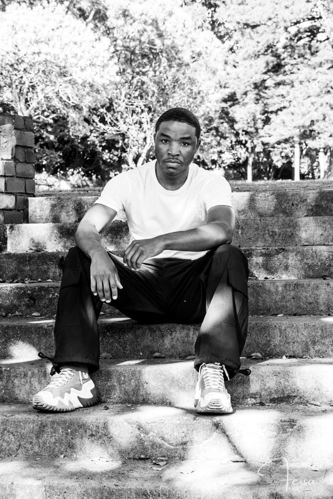 Young person sitting on outdoor steps, wearing a white t-shirt, dark pants, and sneakers, with trees in background.