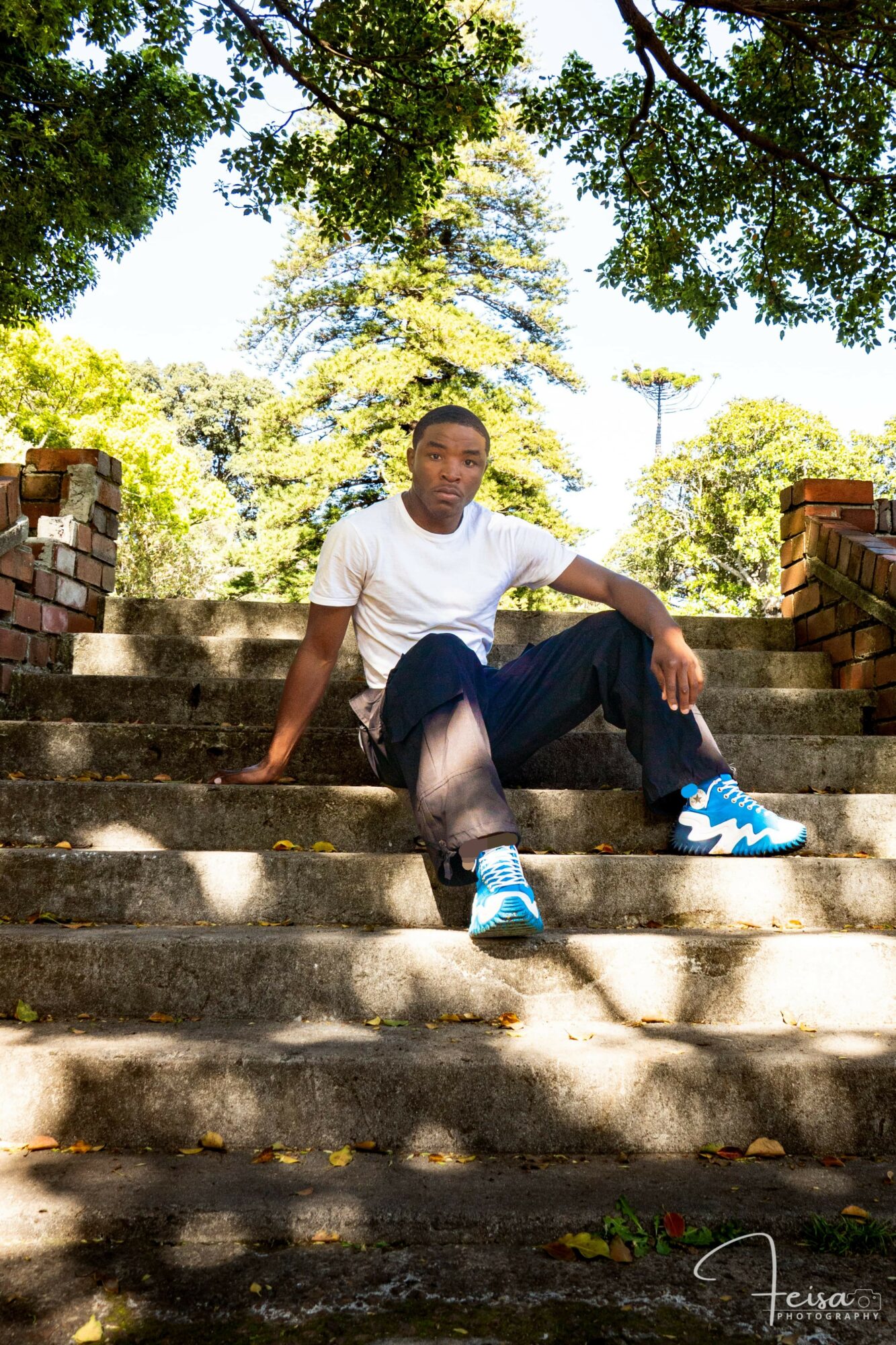Person sitting on outdoor stone steps surrounded by trees and greenery, wearing a white shirt, dark pants, and blue sneakers.