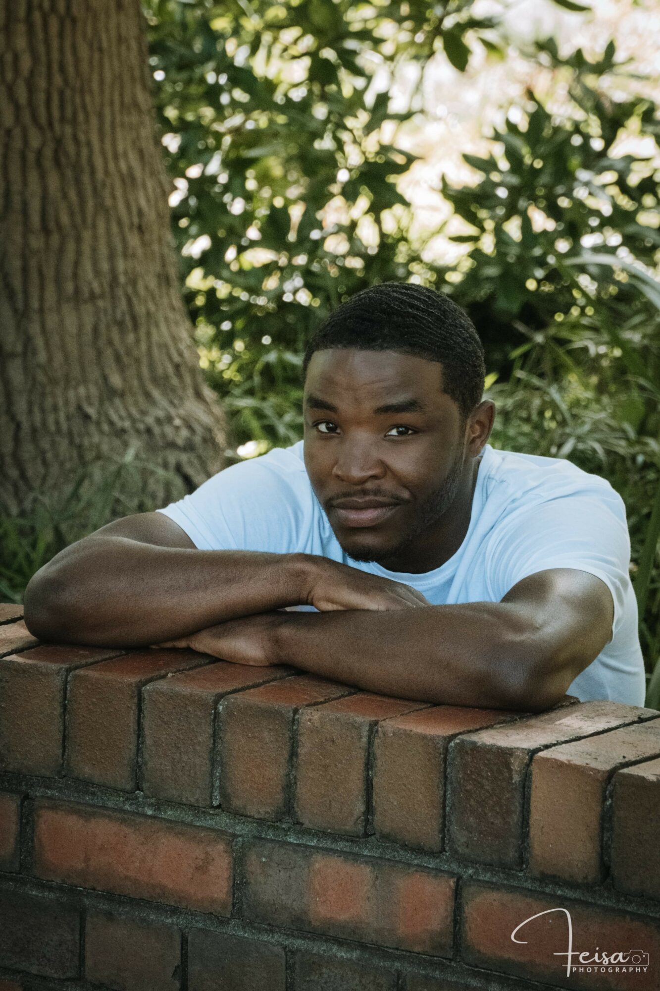 Young man with short hair and a white shirt leans on a brick wall outdoors, with trees and greenery in background.