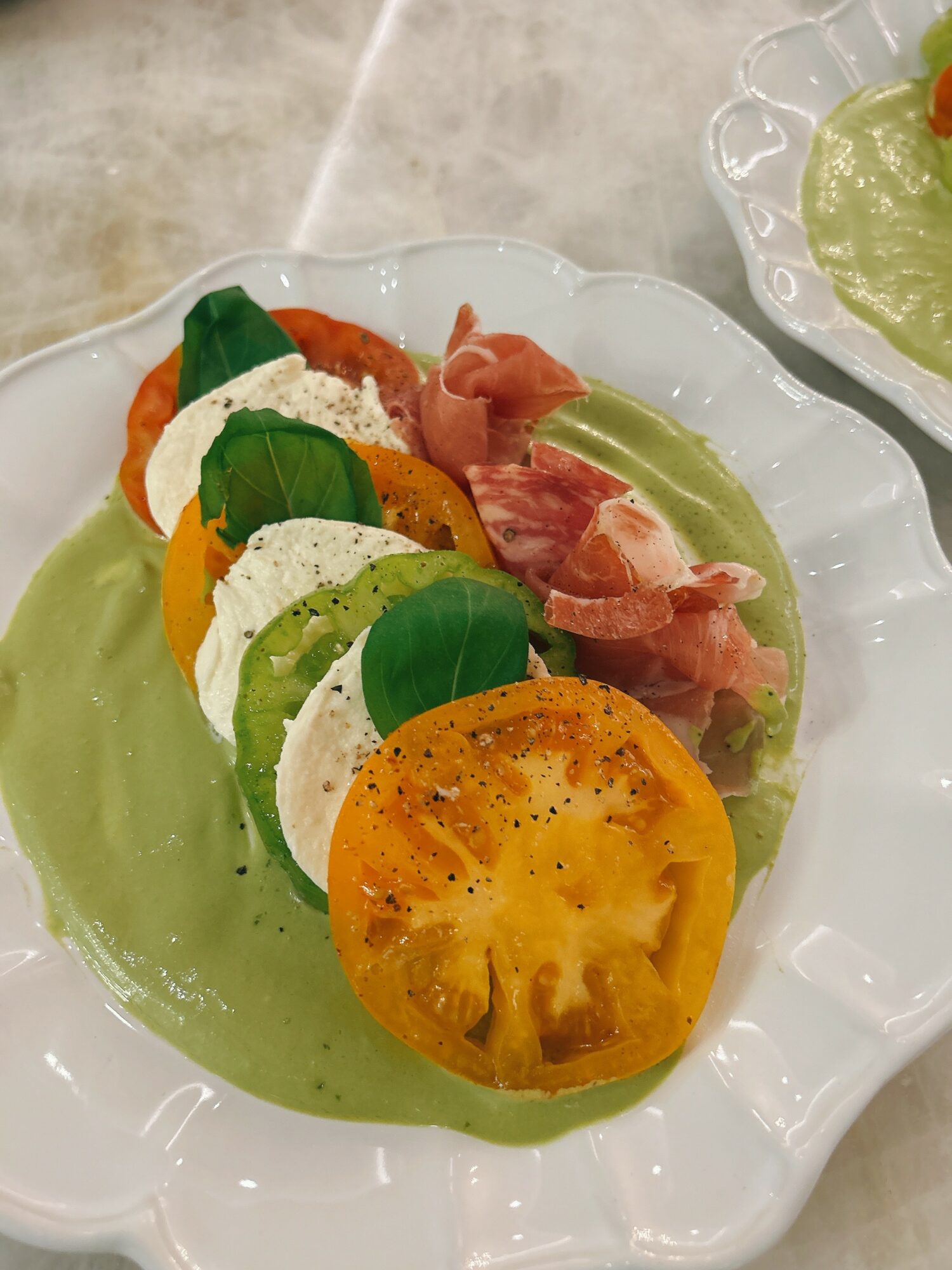 Salad with sliced tomatoes, mozzarella, basil, and prosciutto on a white plate with green sauce.