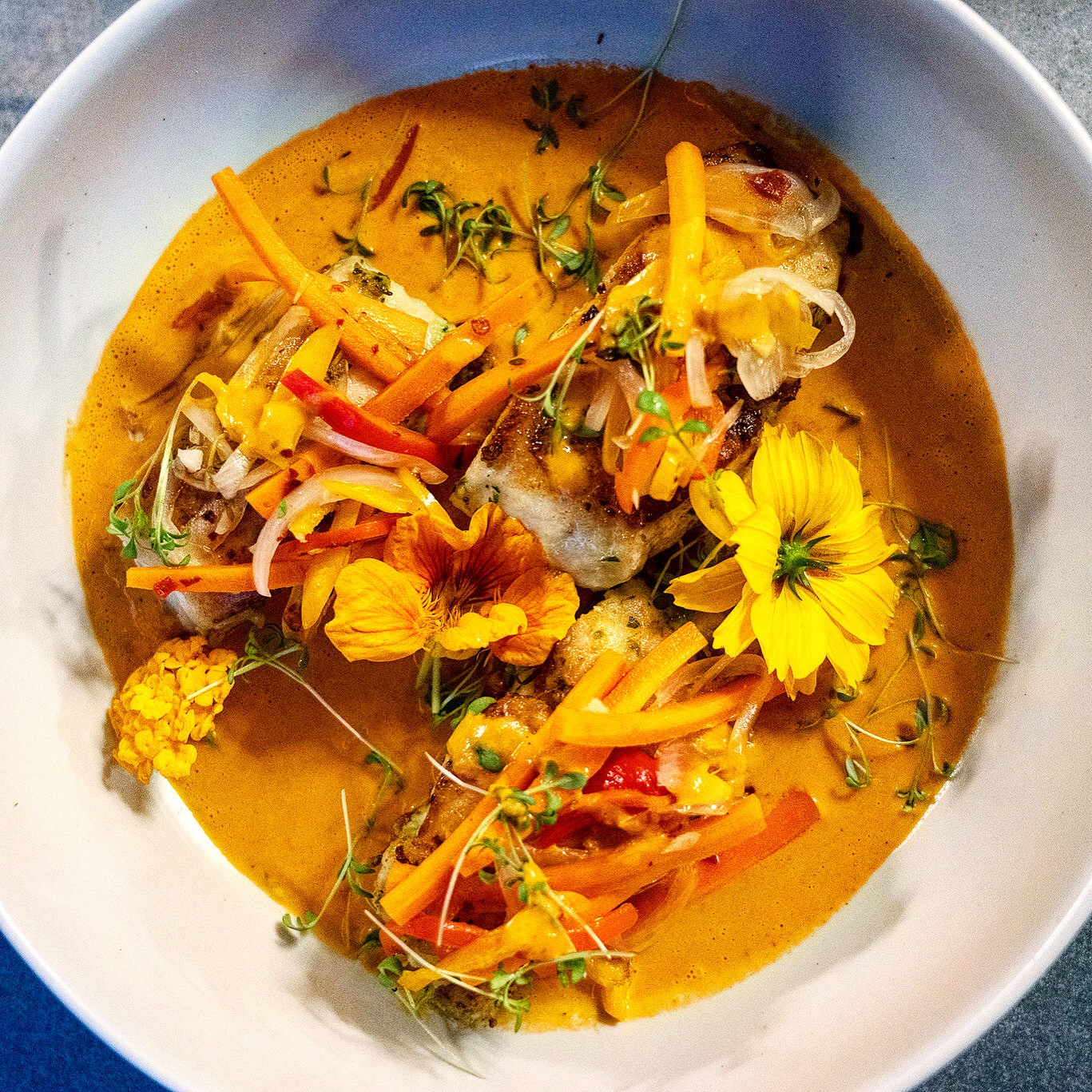 Bowl of colorful curry with vegetables and herbs, garnished with edible flowers, in a white bowl.