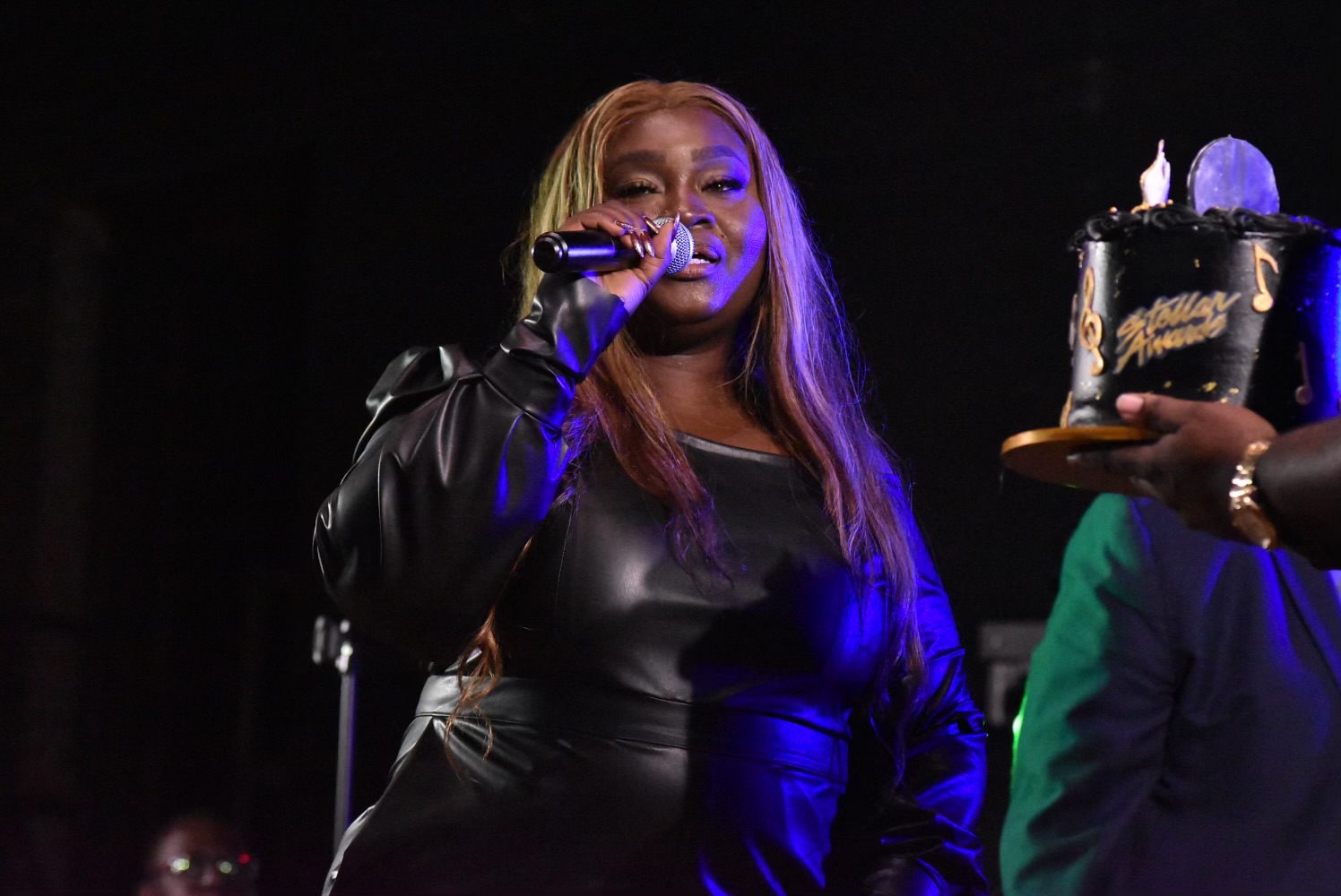 Woman singing into a microphone on stage, holding a cake with lit candles, with a person in green holding the cake.