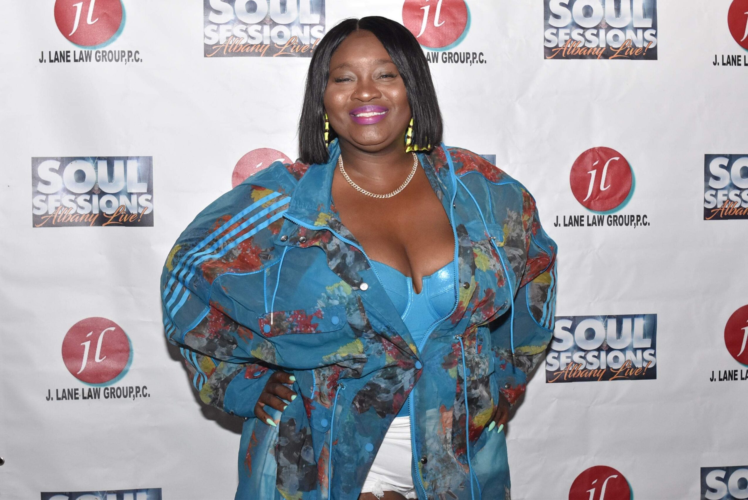 Smiling woman in colorful outfit at event backdrop with logos, posing with hands on hips.