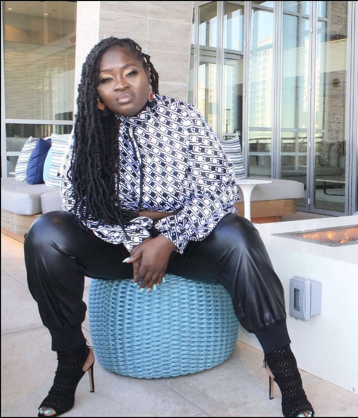 Woman with long braids sitting on a blue round seat in a modern indoor space with large windows.