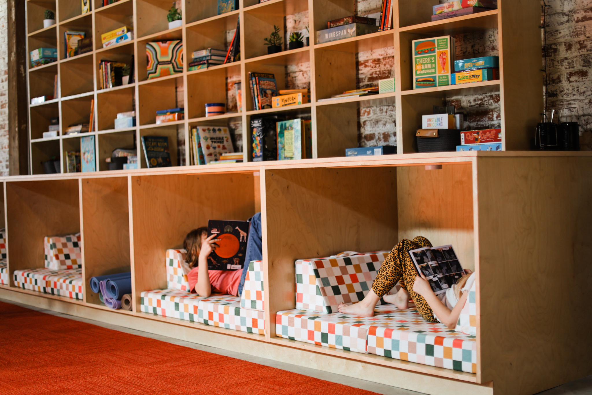 Children are reading books in cubby-style reading nooks with colorful cushions, against a background of shelves filled with books.