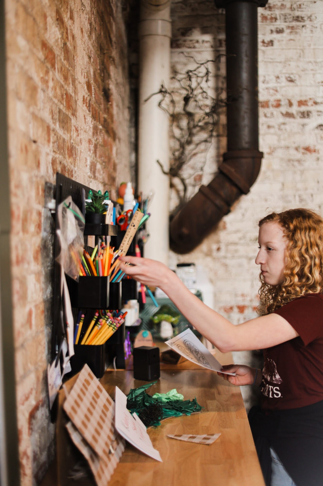 Girl with curly hair reaching for items on a wall-mounted organizer in a brick-walled room.