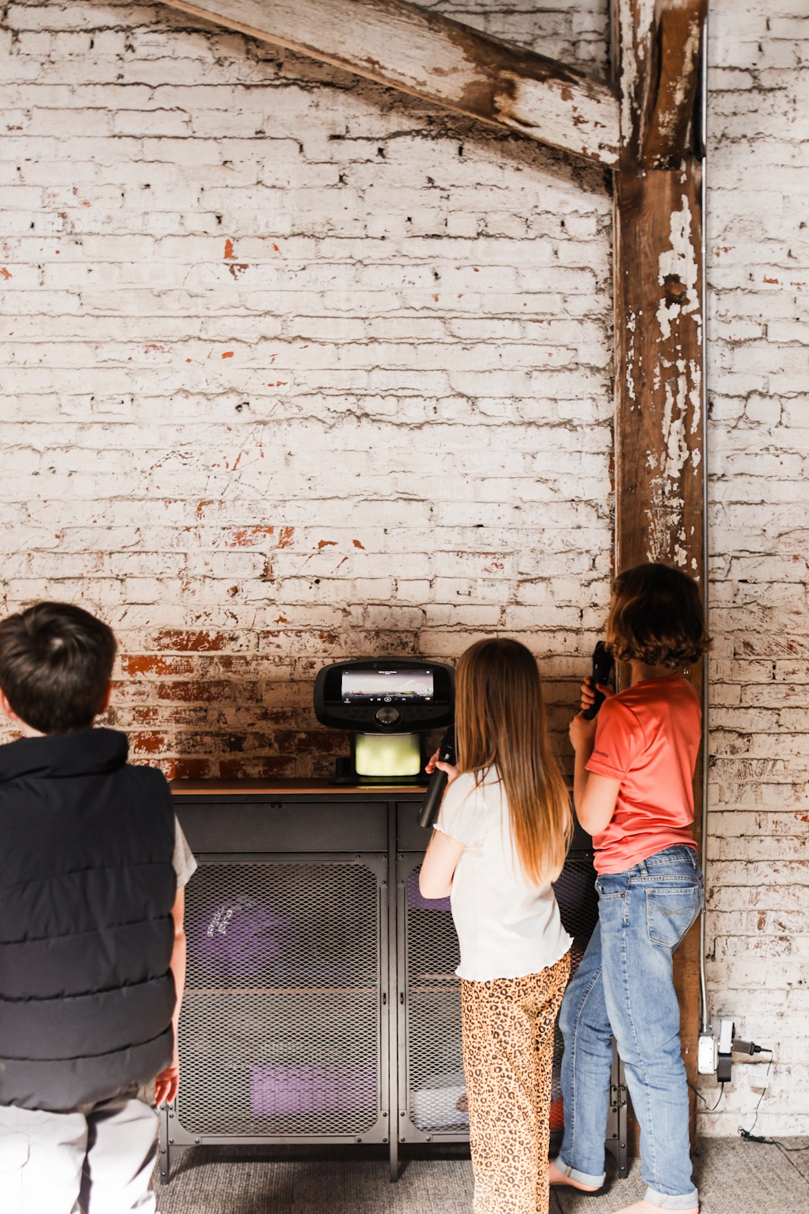 Three children stand in front of a brick wall and a small television, watching it together.
