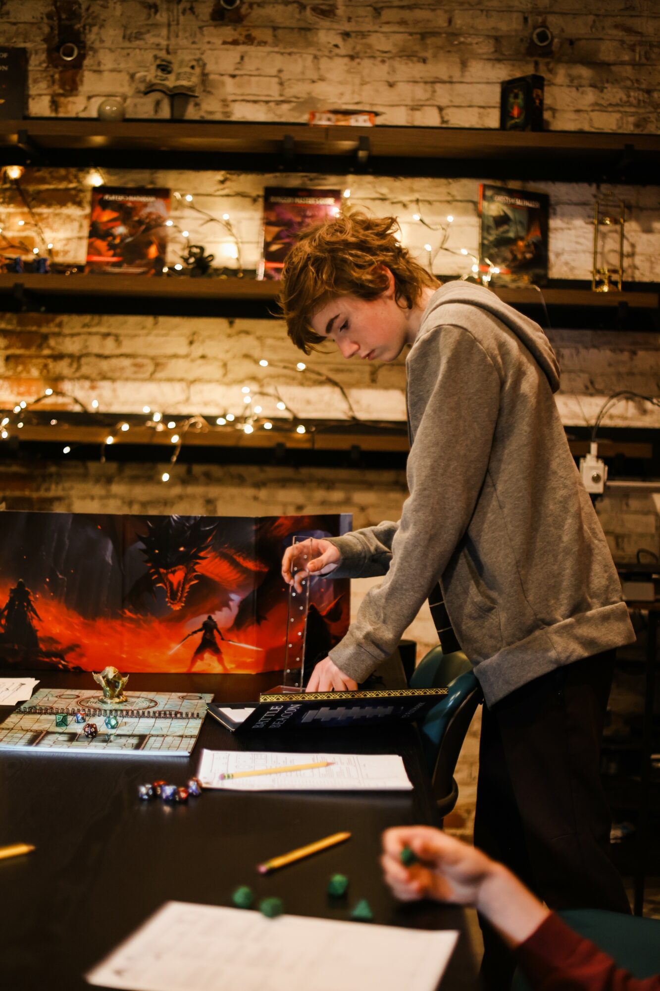 Young person with curly hair in a gray hoodie working at a table in a room with brick walls and shelves, colorful artwork, and string lights.