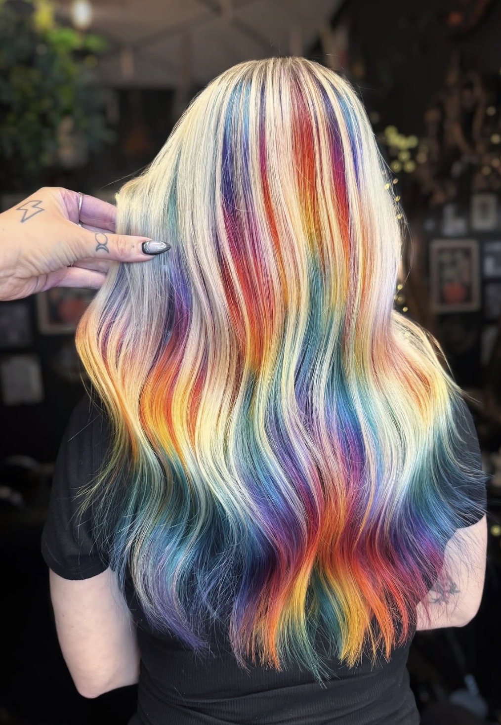 Back of a person with long, wavy, rainbow-colored hair, with a hand pointing at the hair, in an indoor setting.