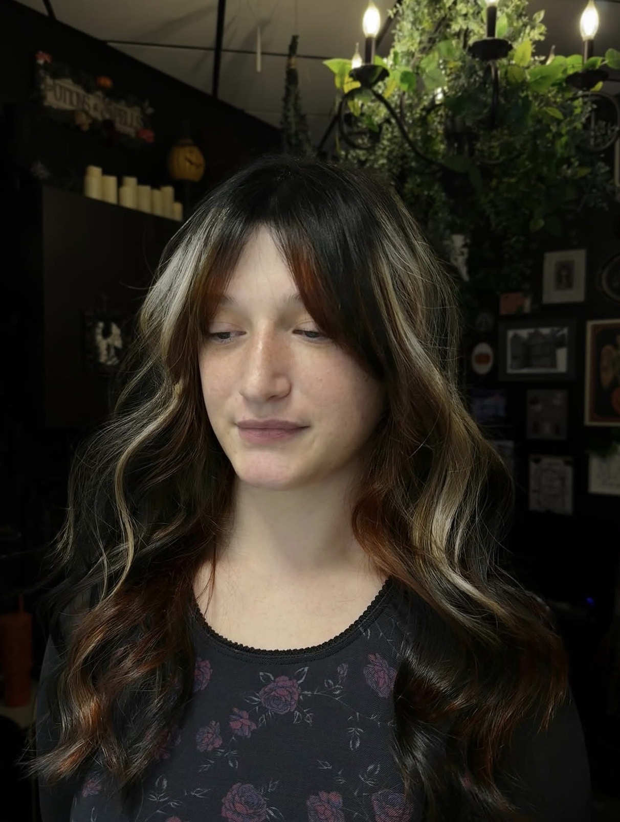 Young woman with long wavy hair and bangs, eyes closed, in a dimly lit room with candles and framed pictures.