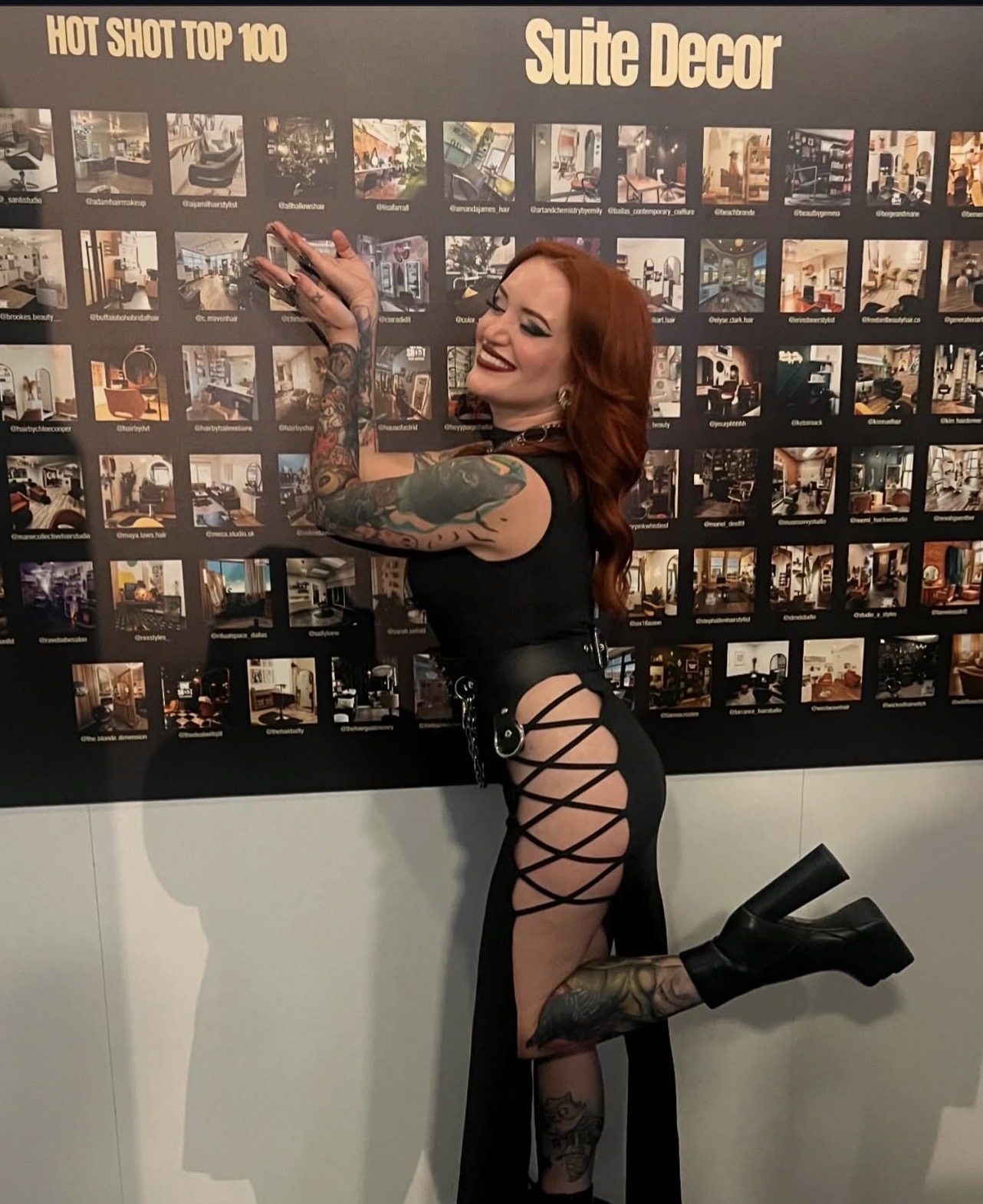 Woman with tattoos and red hair smiling, standing sideways, reaching towards a photo display wall, wearing a black dress with side cutouts, black boots, and holding a phone.