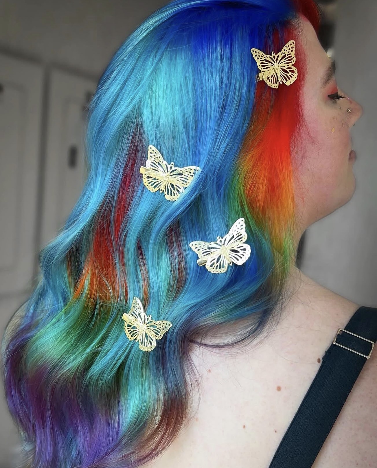 Side view of a person with rainbow-colored hair and butterfly hair clips, hair flowing over shoulder.