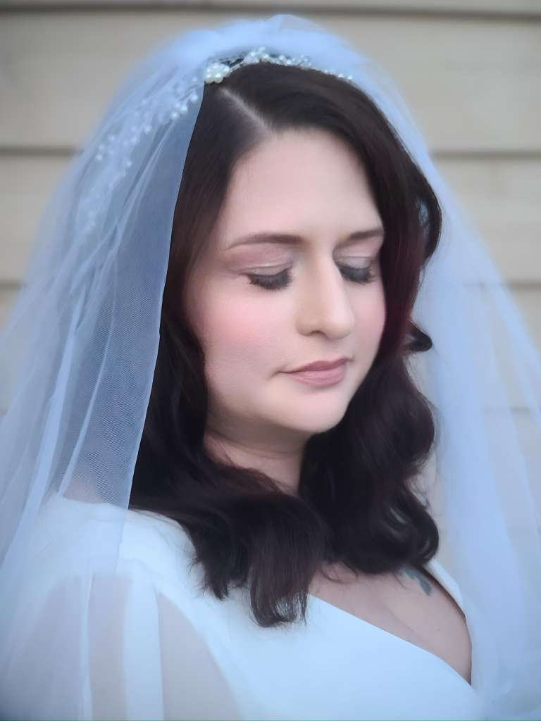 Woman with dark hair and closed eyes wearing a white dress and a light blue veil, outdoors.