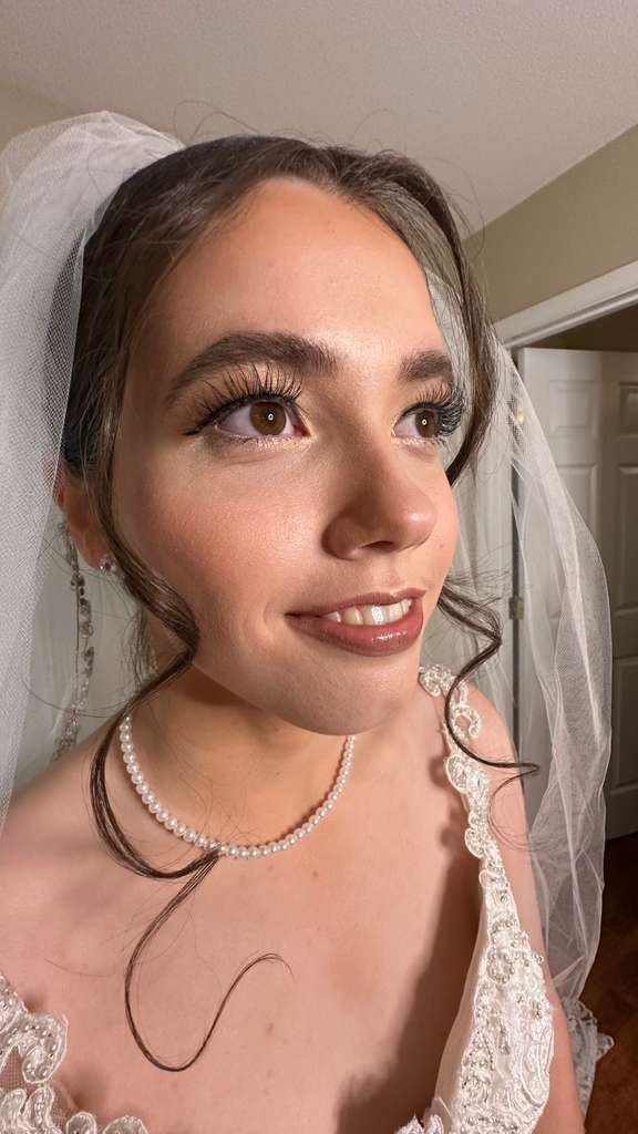 Close-up of a woman with makeup, wearing a pearl necklace and lace dress, smiling, with a veil in her hair.