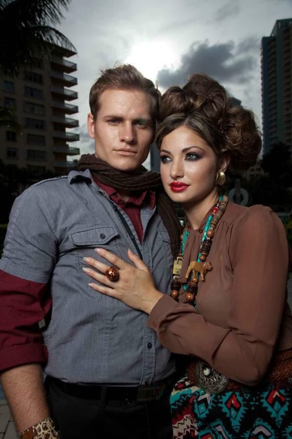 A man and woman pose outdoors with buildings and cloudy sky in background. The woman has curly hair and colorful jewelry.