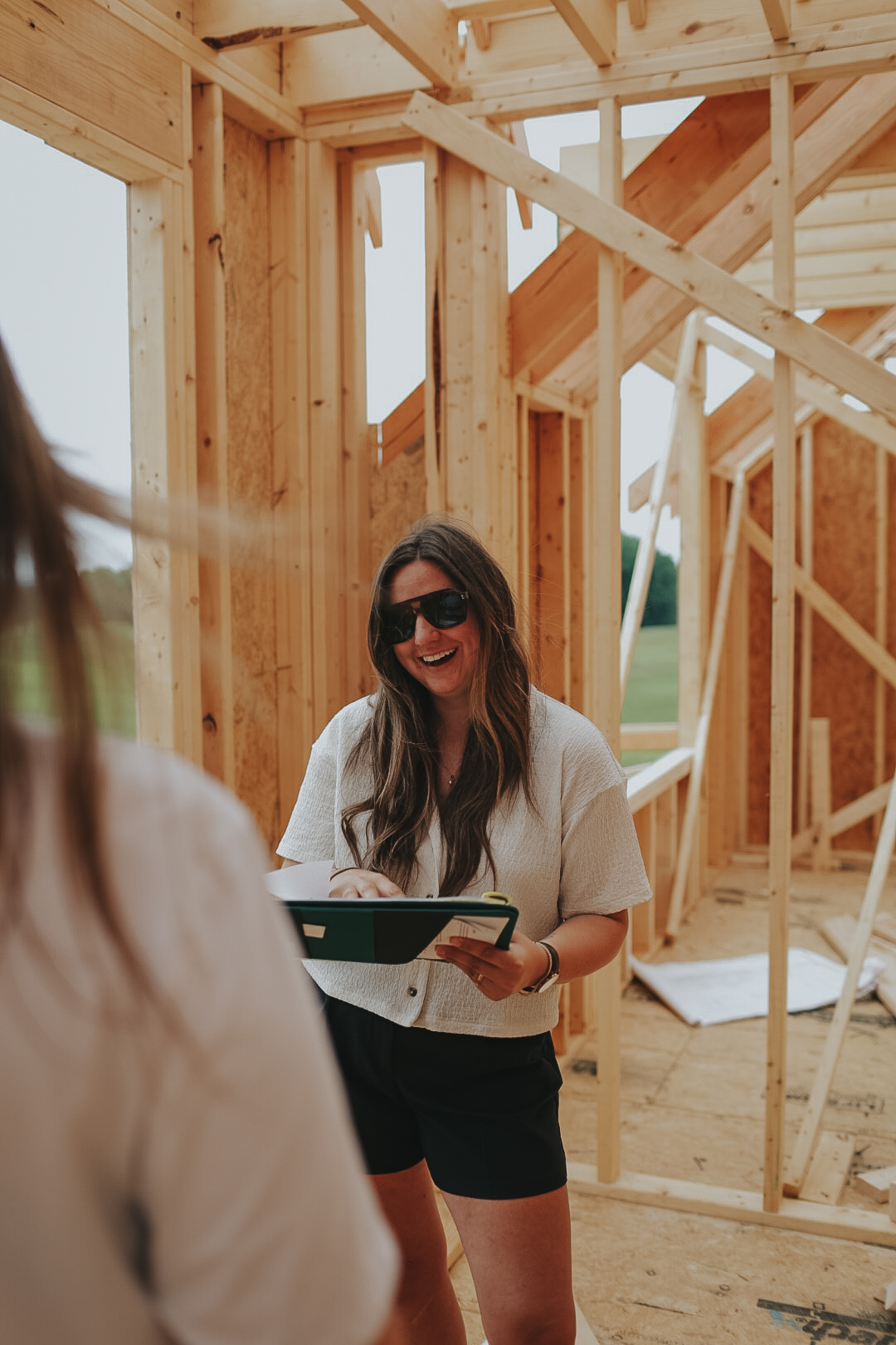 Woman with long dark hair and sunglasses smiling at a construction site, holding a tablet, with wooden framing in background.
