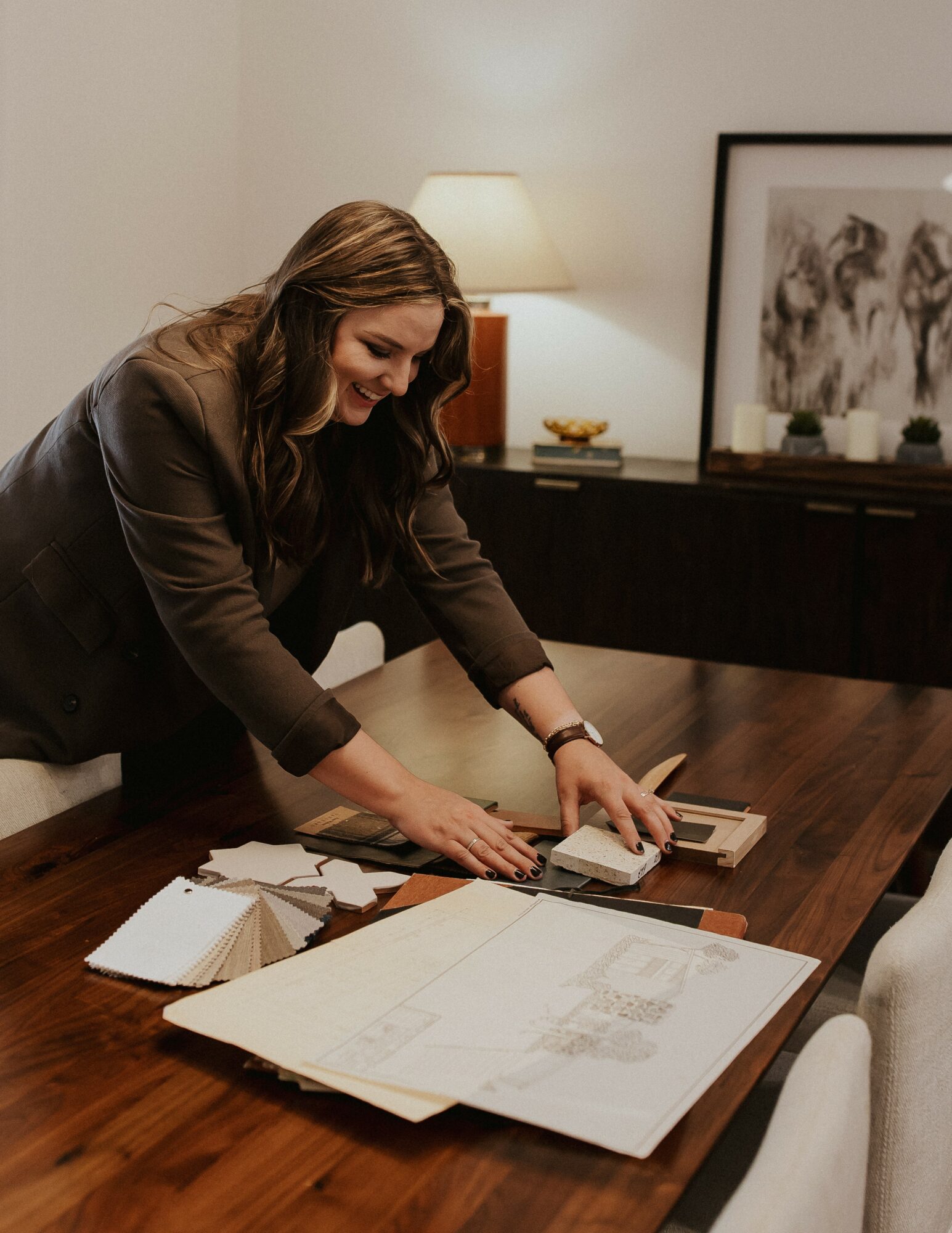 Woman leaning over a table, working with architectural plans and drawings, in a room with a lamp and framed artwork.