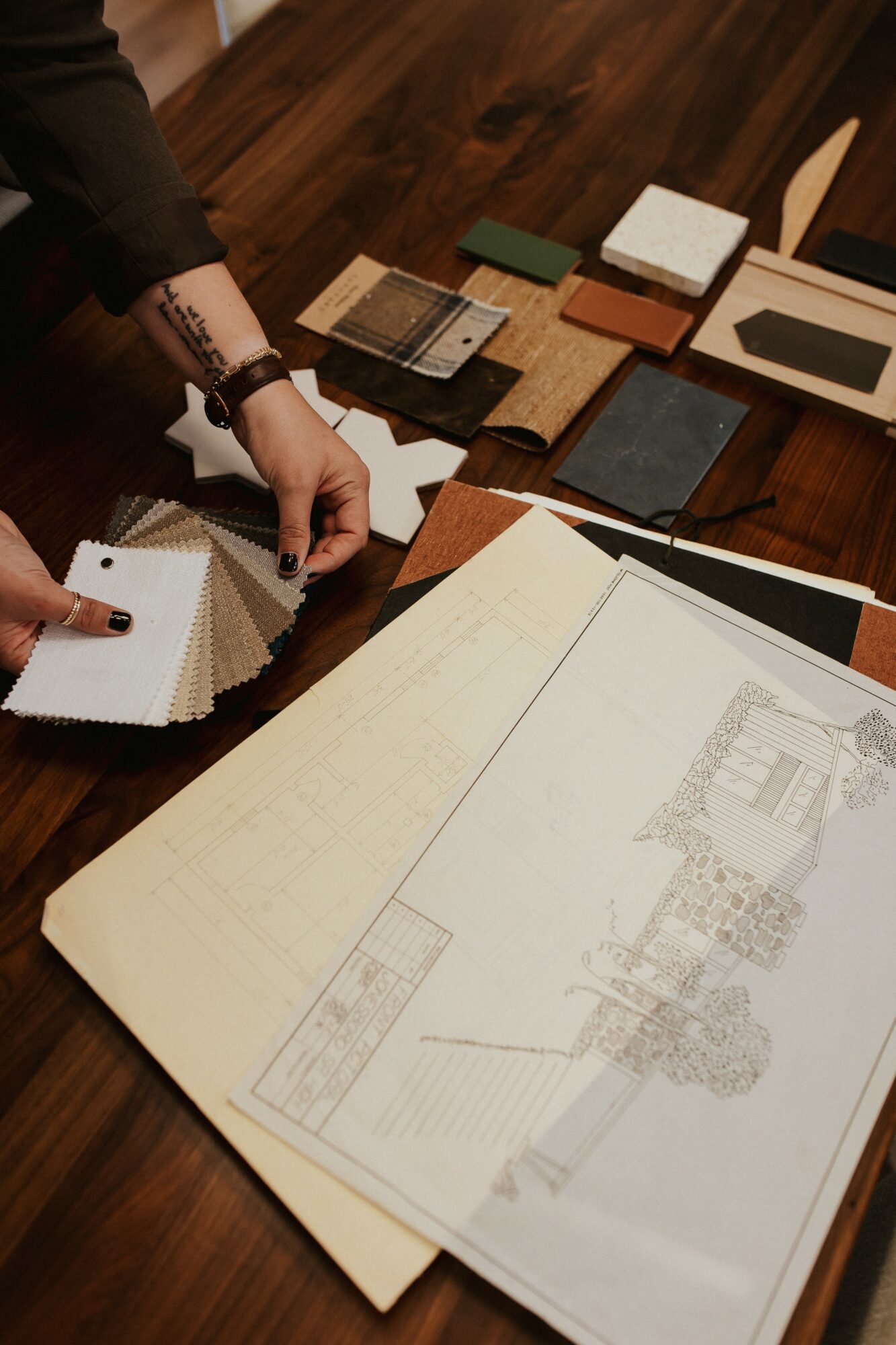 Person's hand holding fabric samples over a wooden table with design plans and color swatches.