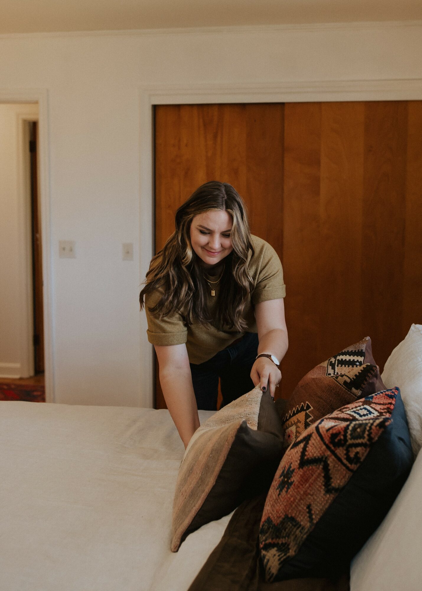 Woman arranging pillows on a bed in a room with wooden closet doors, smiling, with beige walls.