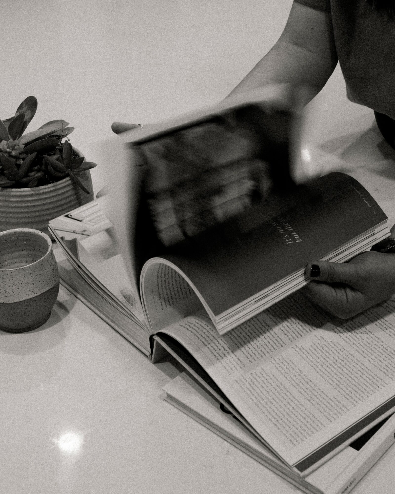 Person flipping through a magazine on a table with a potted plant and a cup nearby.