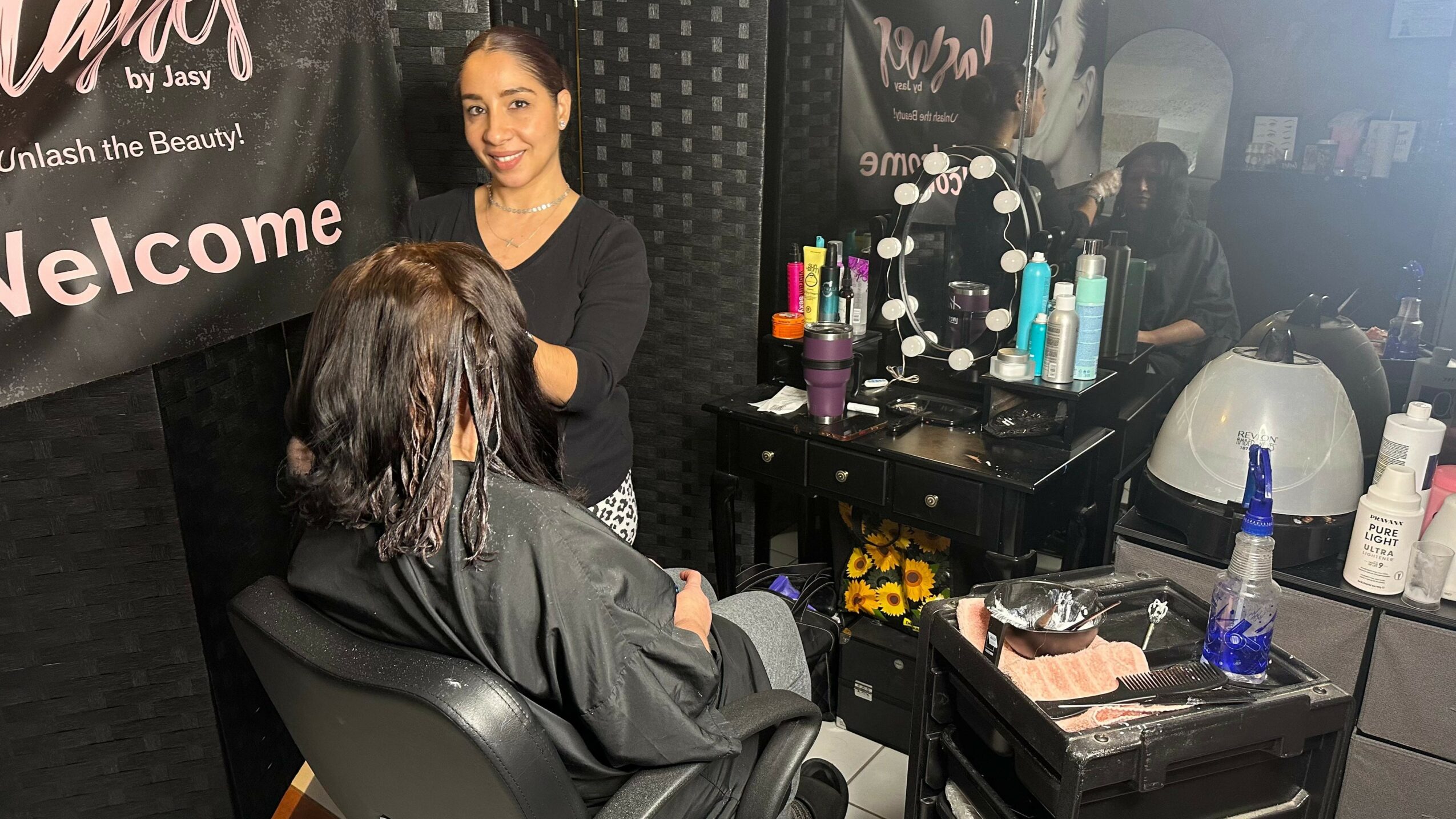 A woman with dark hair sits in a salon chair, facing a stylist with long dark hair, in a beauty salon. The background has beauty products and a welcome sign.