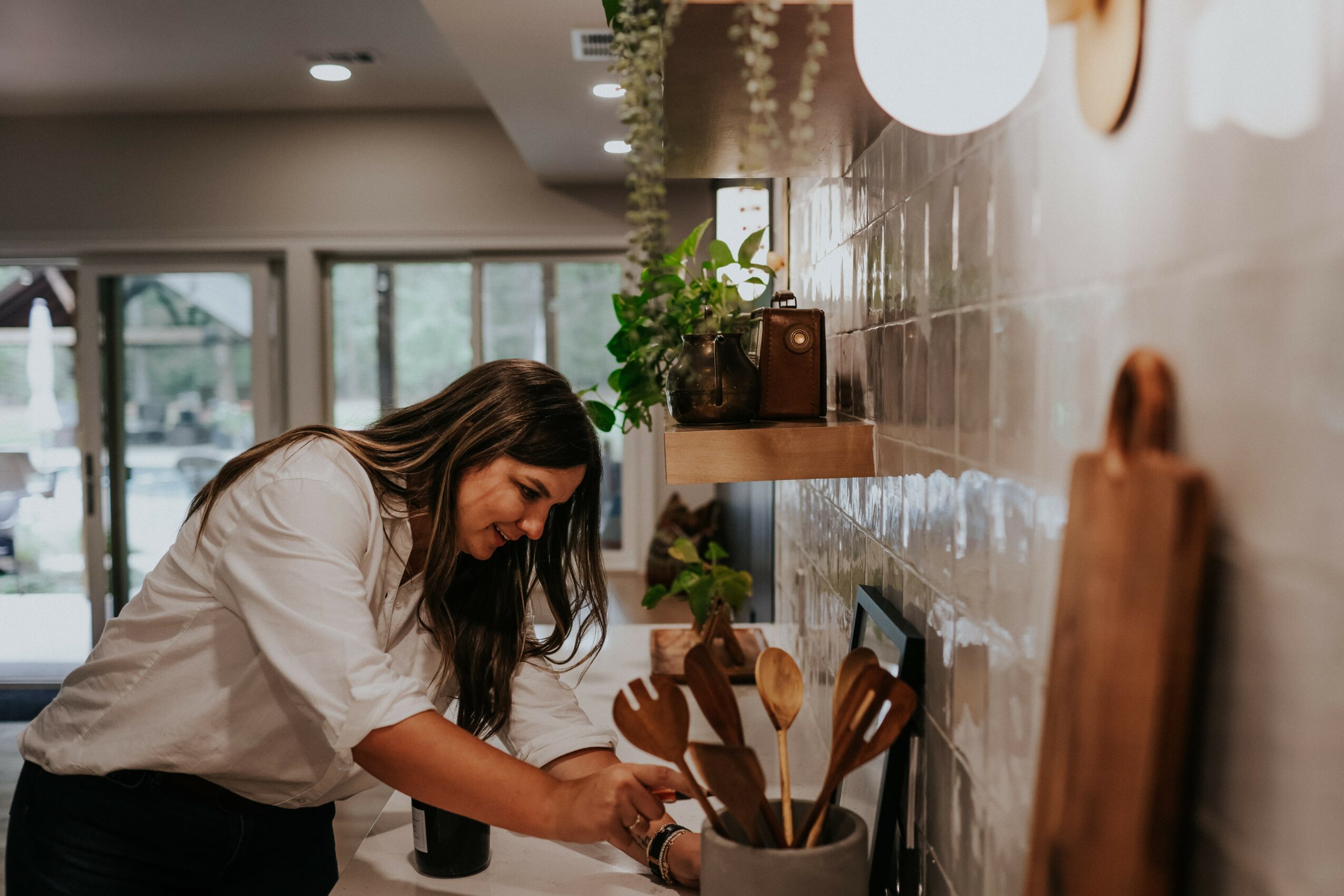 Woman leaning over a counter in a modern cafe, smiling, with kitchen utensils and plants on shelves behind her.