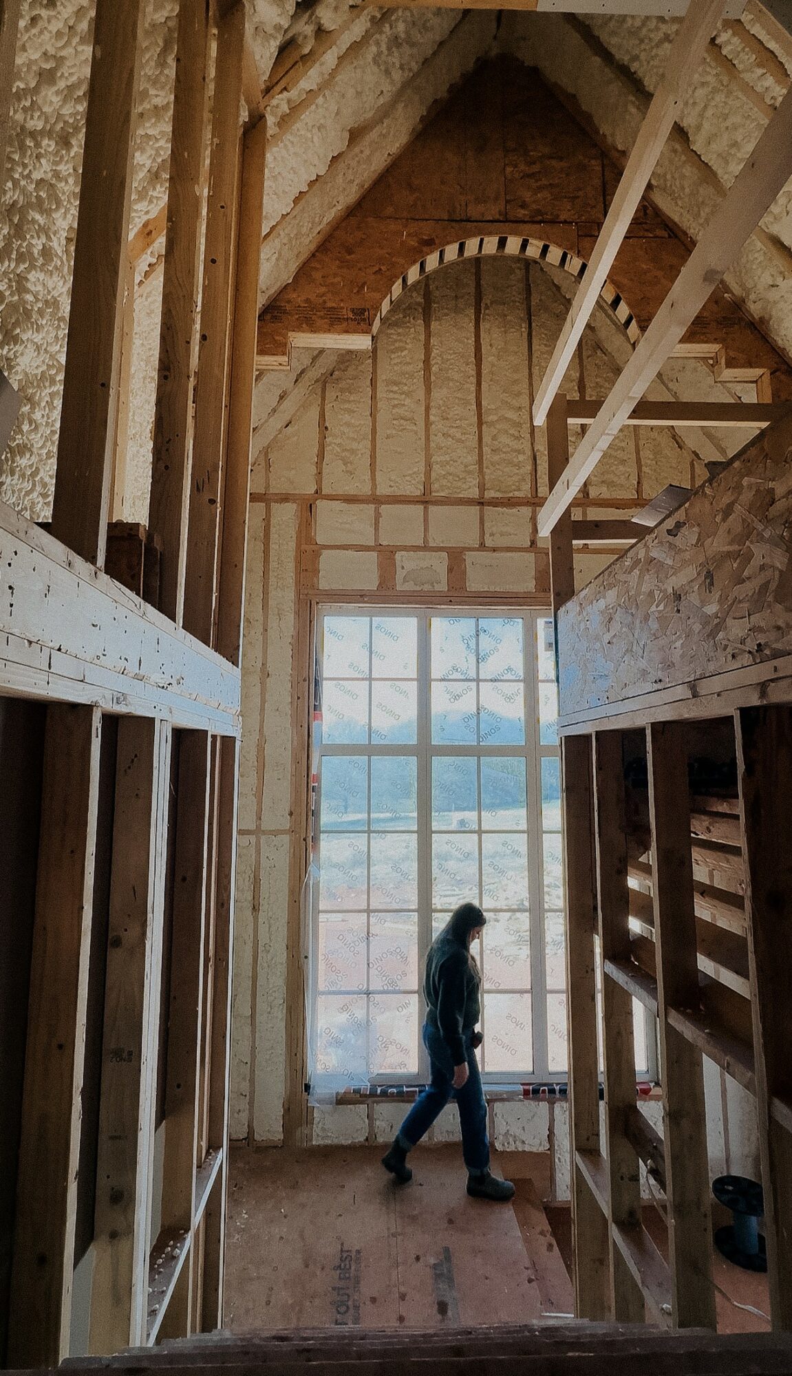 Person walking inside a tall, unfinished brick building with large window and wooden framing, sunlight streaming through window.