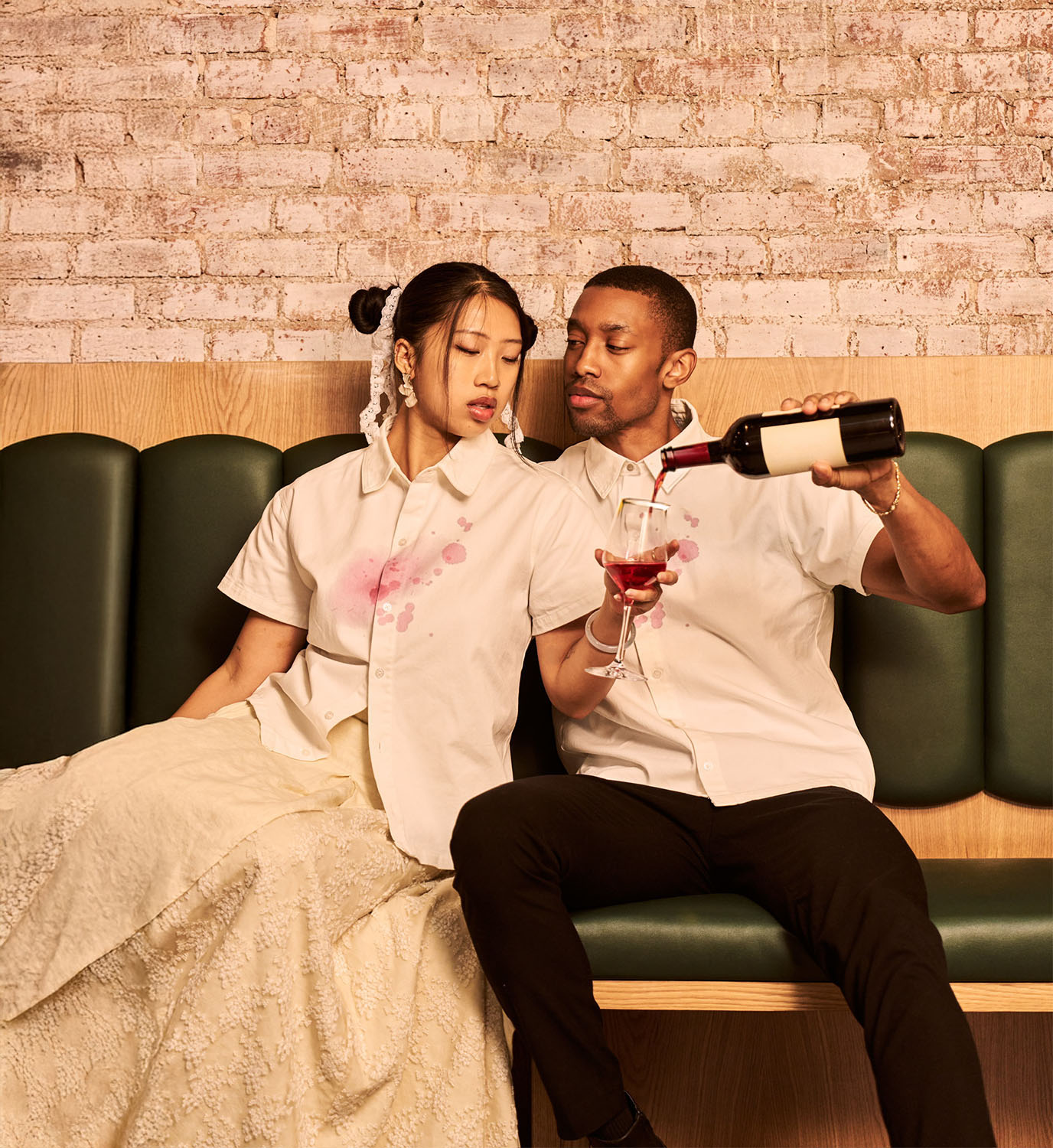 Two people sitting on a green bench against a brick wall, one pouring wine into the other's glass, both dressed casually.