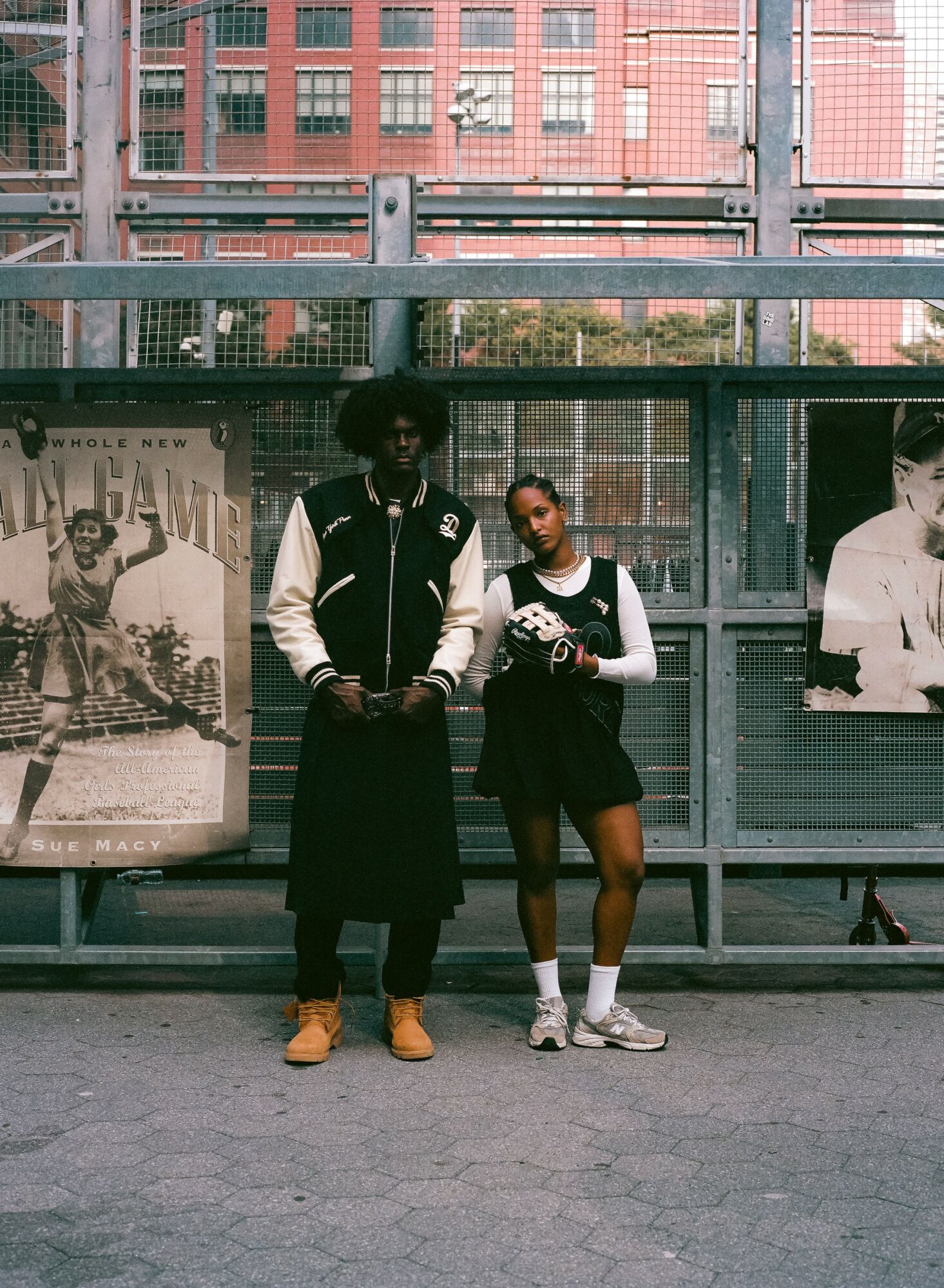 Two people standing in front of a bus stop with posters, one wearing a jacket and the other in a dress, both smiling.