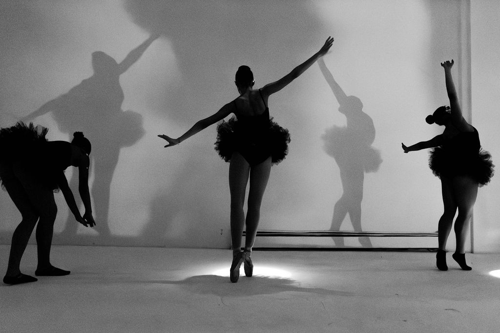 Five ballet dancers perform on stage with shadows cast on the wall behind them, some in poses and others adjusting costumes.