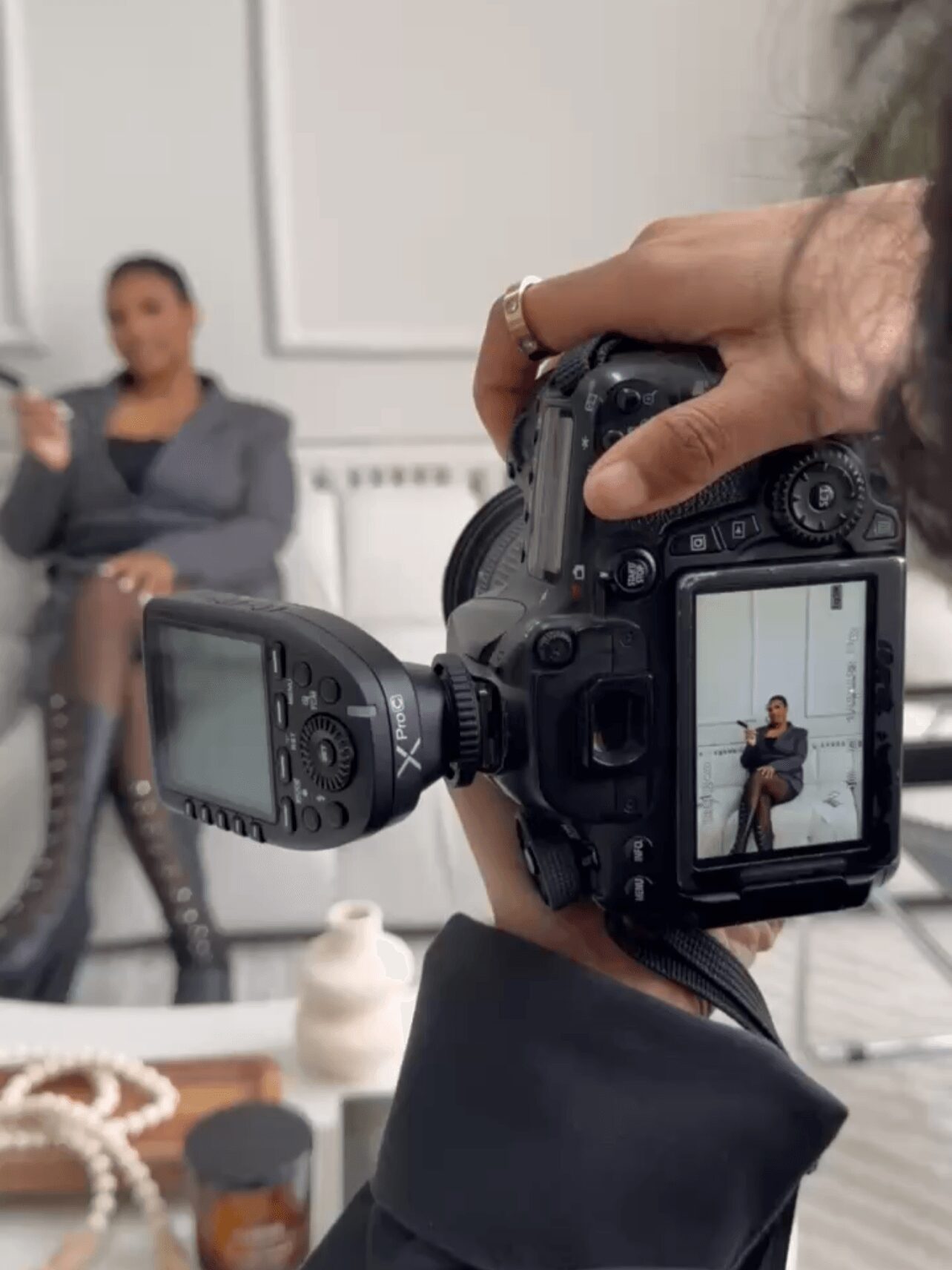 Photographer taking a picture of a woman sitting on a chair in a room, camera screen showing the subject, with a blurred background.