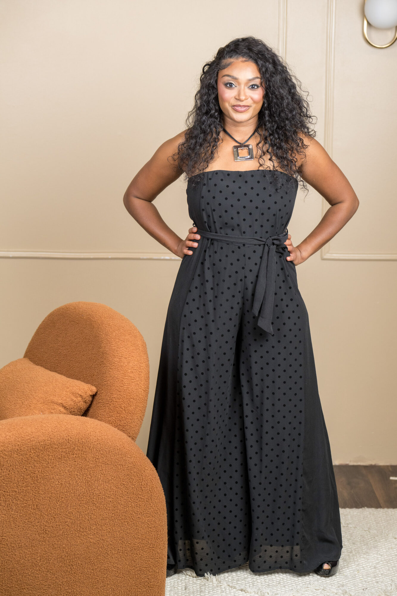 Woman with curly hair in a strapless black polka dot dress standing indoors with hands on hips.