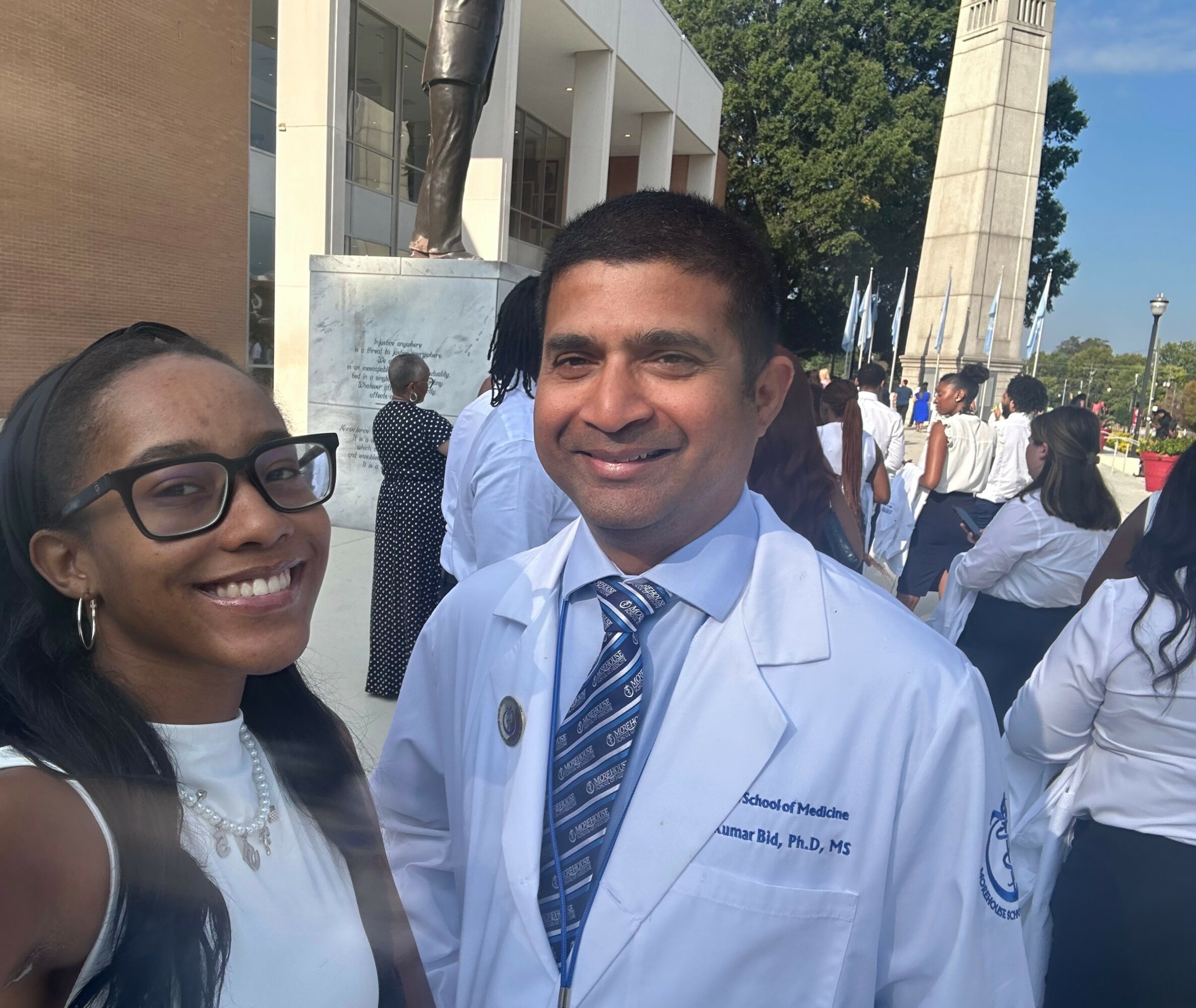 Two people smiling outdoors, one in a white coat and tie, the other in glasses and a white top, with a crowd and monument in background.