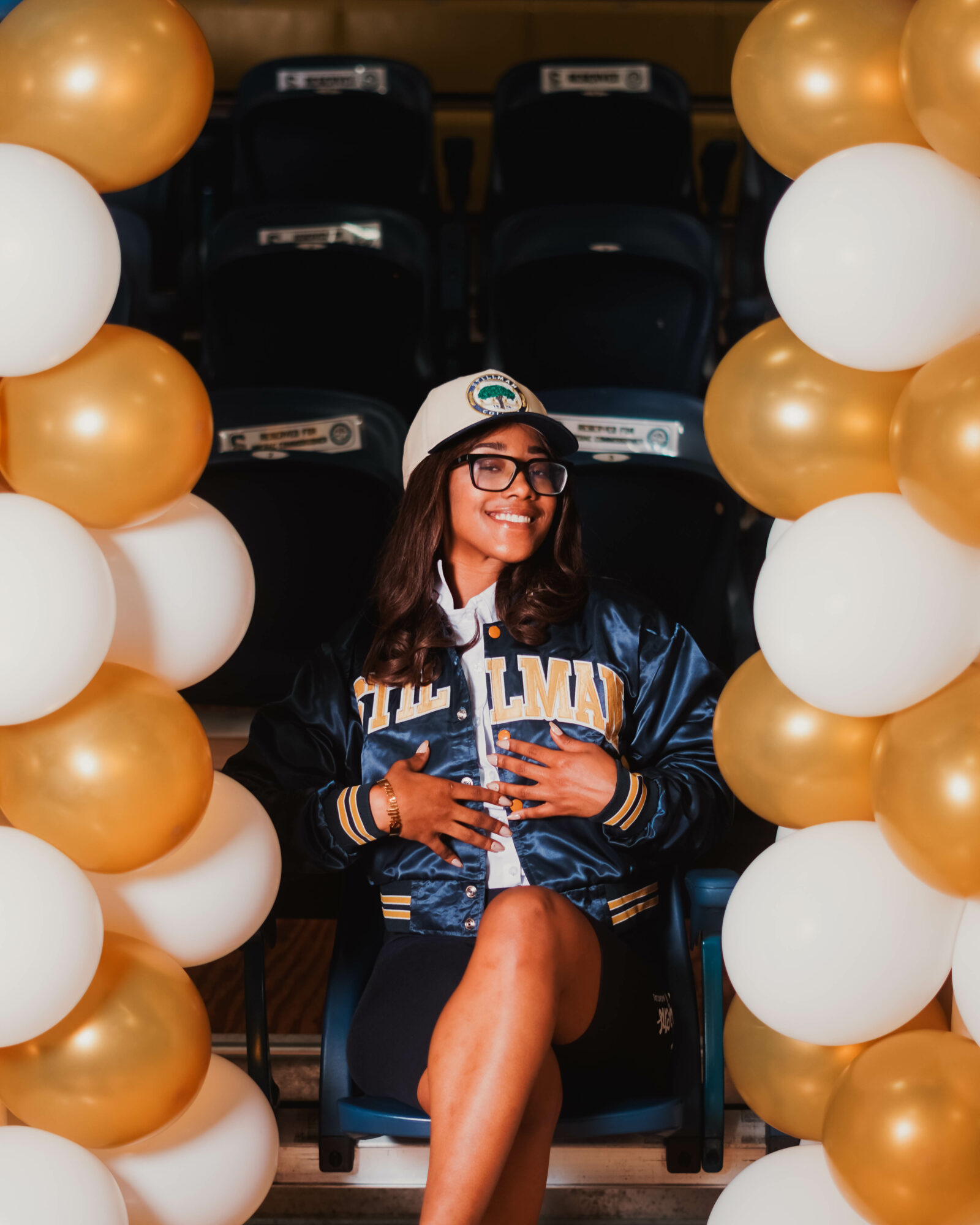 Young woman smiling, sitting between gold and white balloons, wearing glasses, a cap, and a blue jacket, in a stadium.