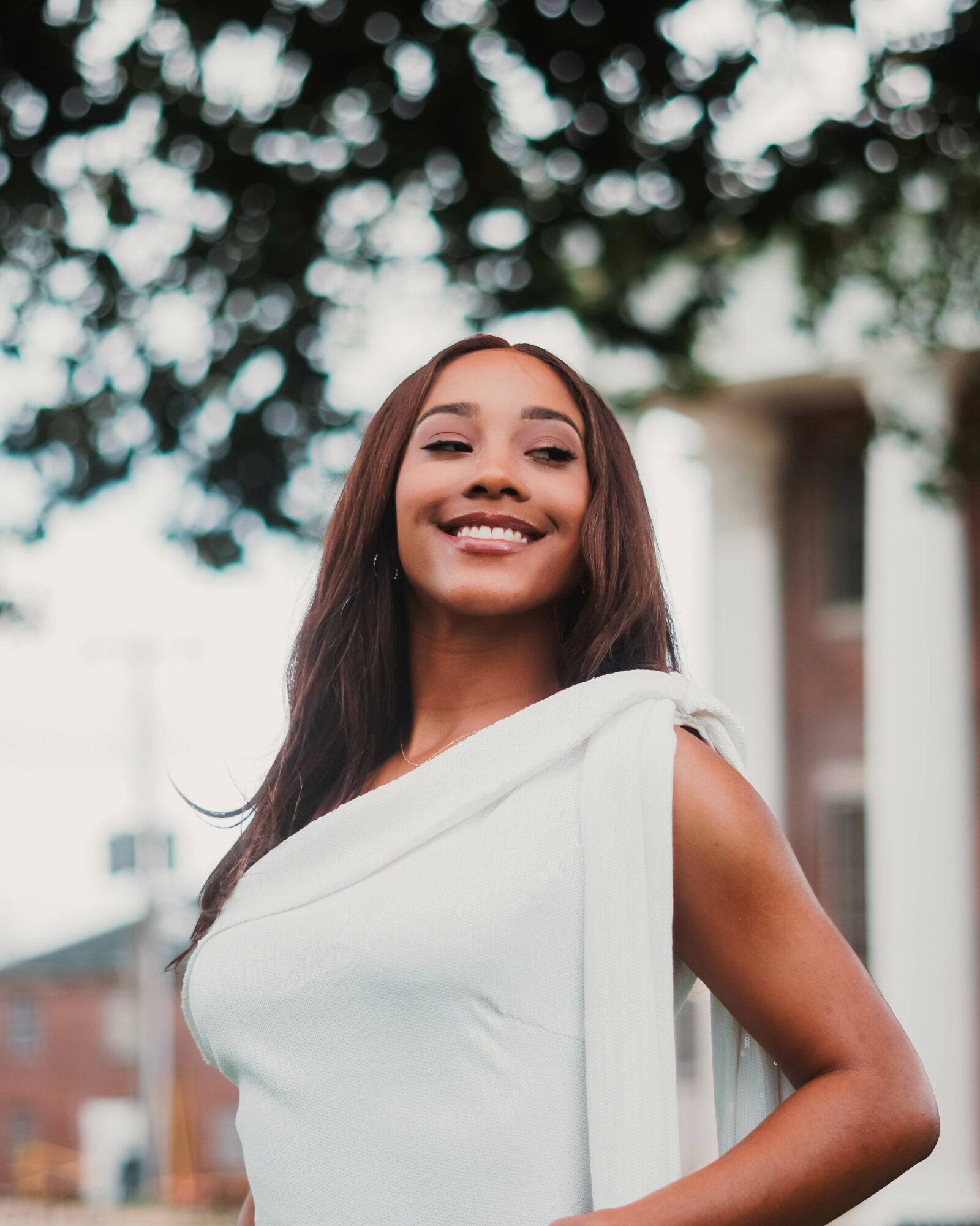 Smiling woman with long dark hair wearing a white dress outdoors, trees and buildings in background.