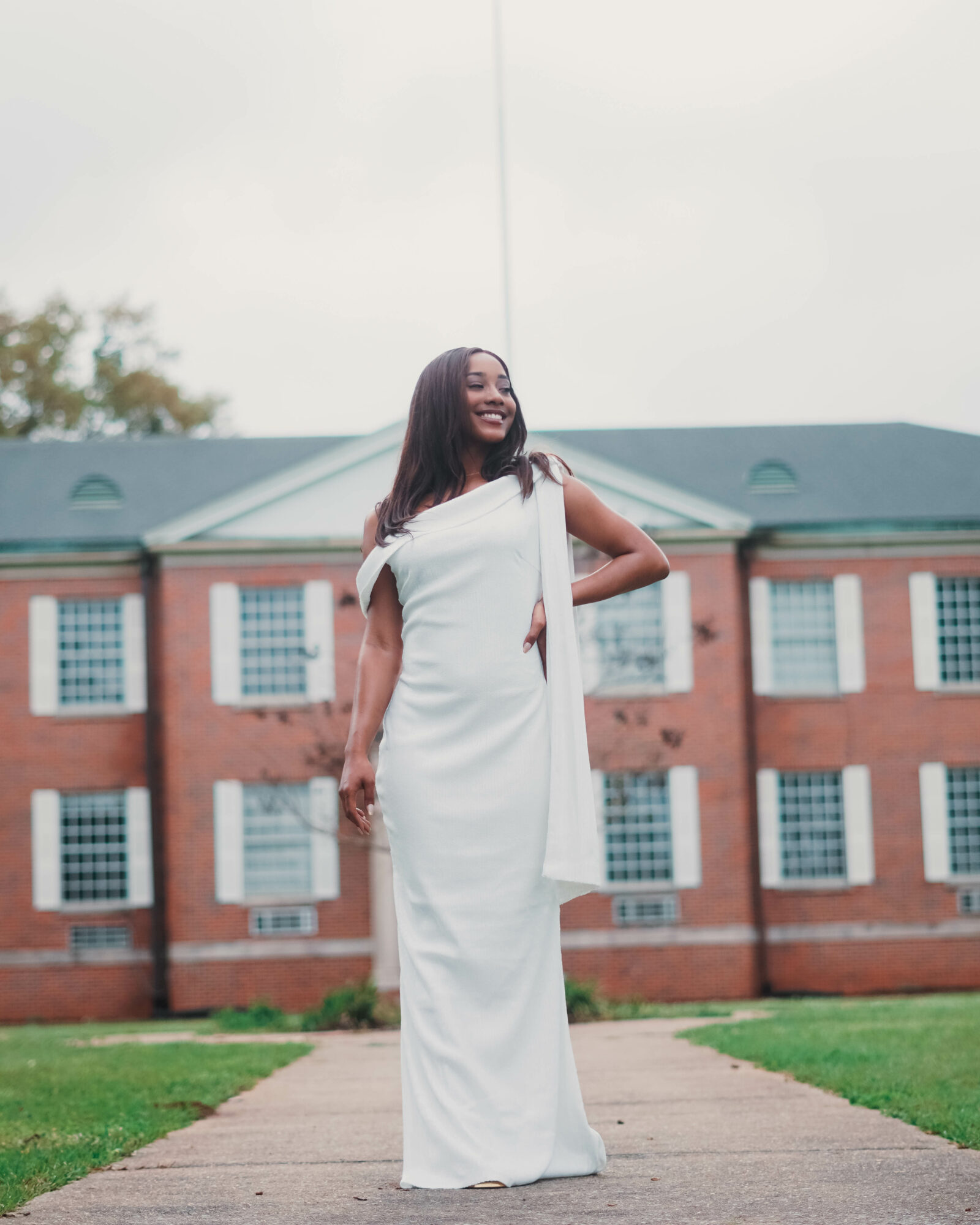 Woman in a white dress standing on a pathway in front of a brick building with multiple windows, smiling.