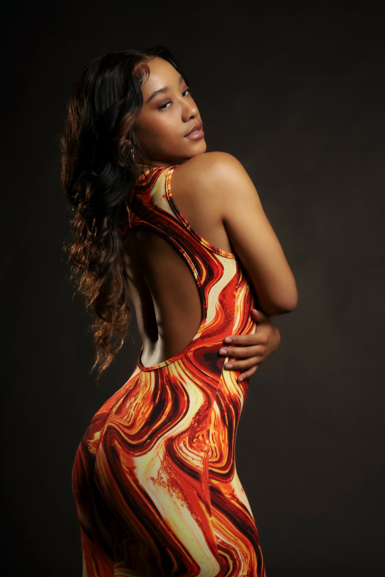 Woman with long wavy hair in a colorful, swirling patterned dress, posing against a dark background.