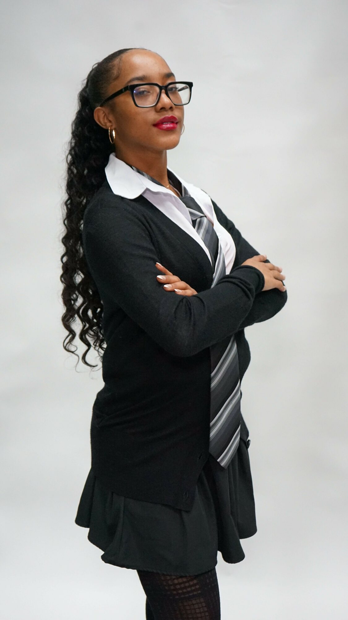 Woman with curly hair and glasses, dressed in professional attire, leaning forward with arms crossed, against a plain background.