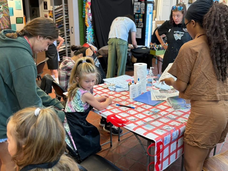 Children and adults gather around a table with a red and white checkered tablecloth, engaging in activities in a room.