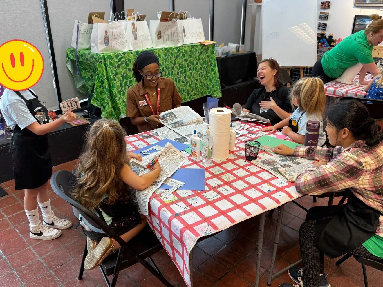 Group of children and an adult sitting around a table with a red and white checkered tablecloth, engaging in activities.